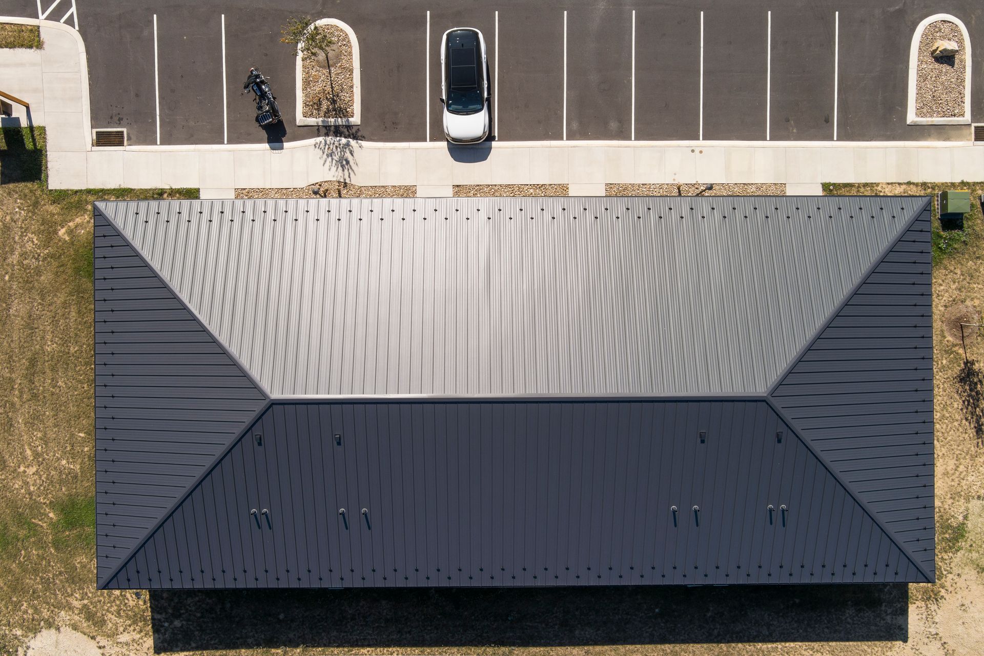 An aerial view of a dark grey, metal-roofed building adjacent to a parking lot containing one car and a motorcycle.