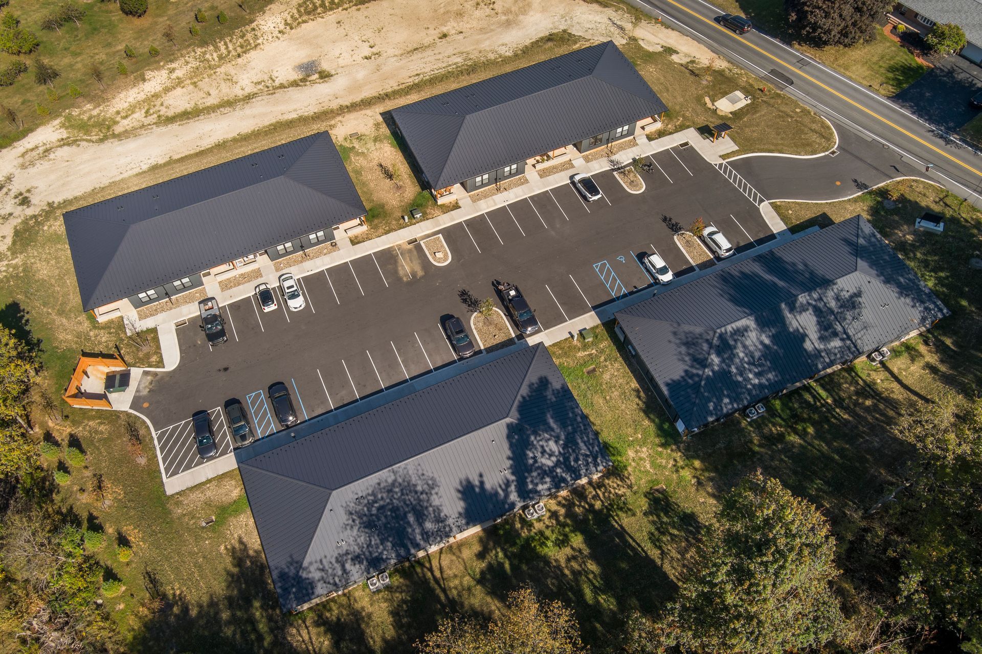 An aerial view of four dark metal-roofed buildings arranged around a central paved parking lot in a rural, wooded setting.
