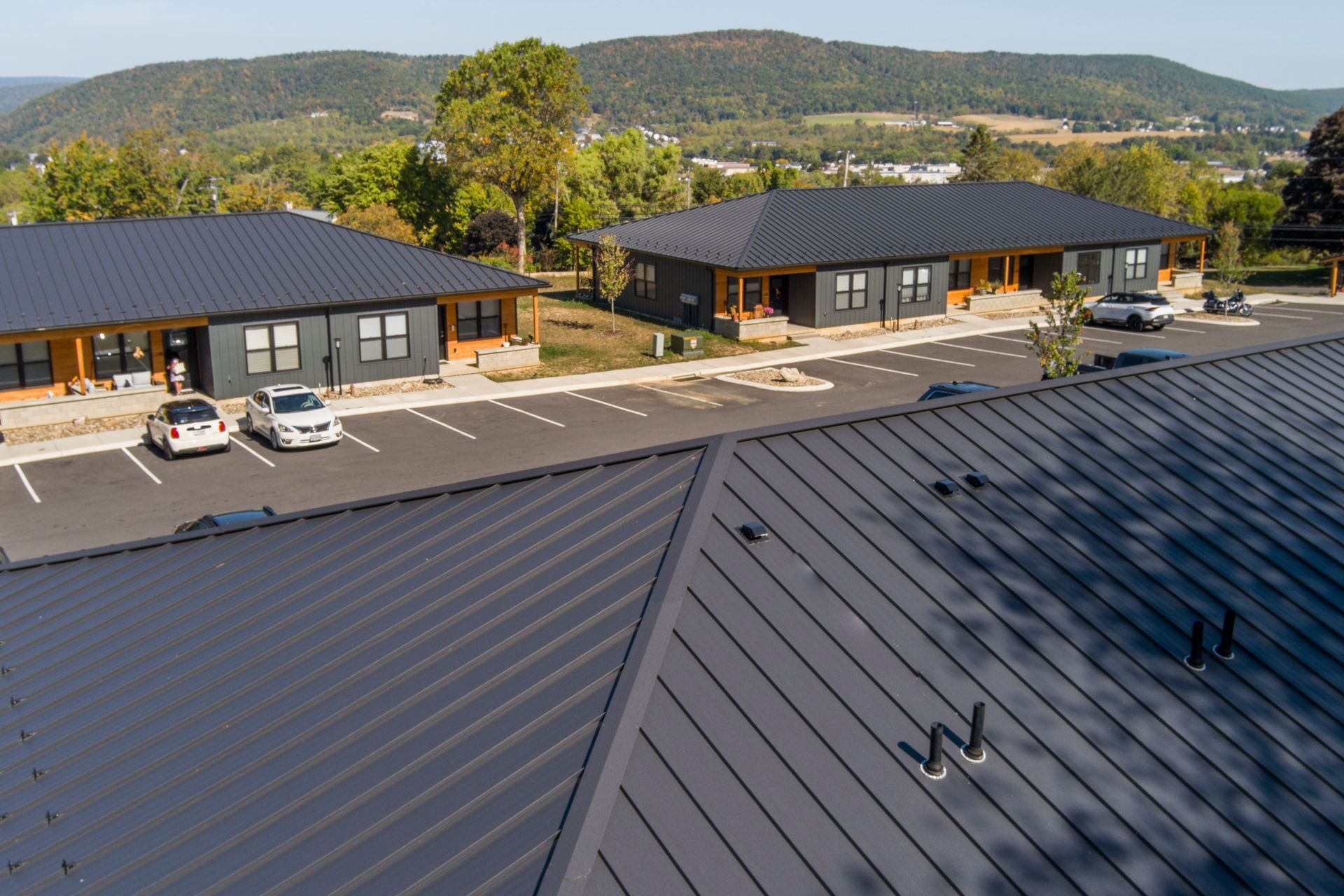 High-angle view of modern, dark metal-roofed buildings in a parking lot surrounded by trees and mountains.