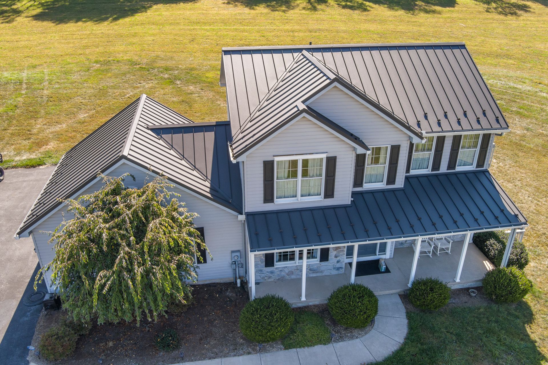Aerial view of a two-story gray house with a dark metal roof, front porch, and surrounding lawn on a sunny day.