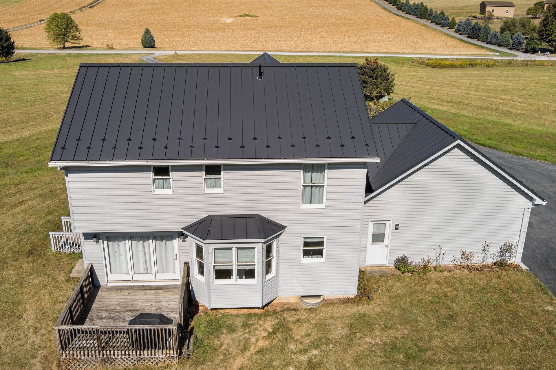 An aerial view of a two-story gray house with a new black metal roof, surrounded by rural, grassy fields.