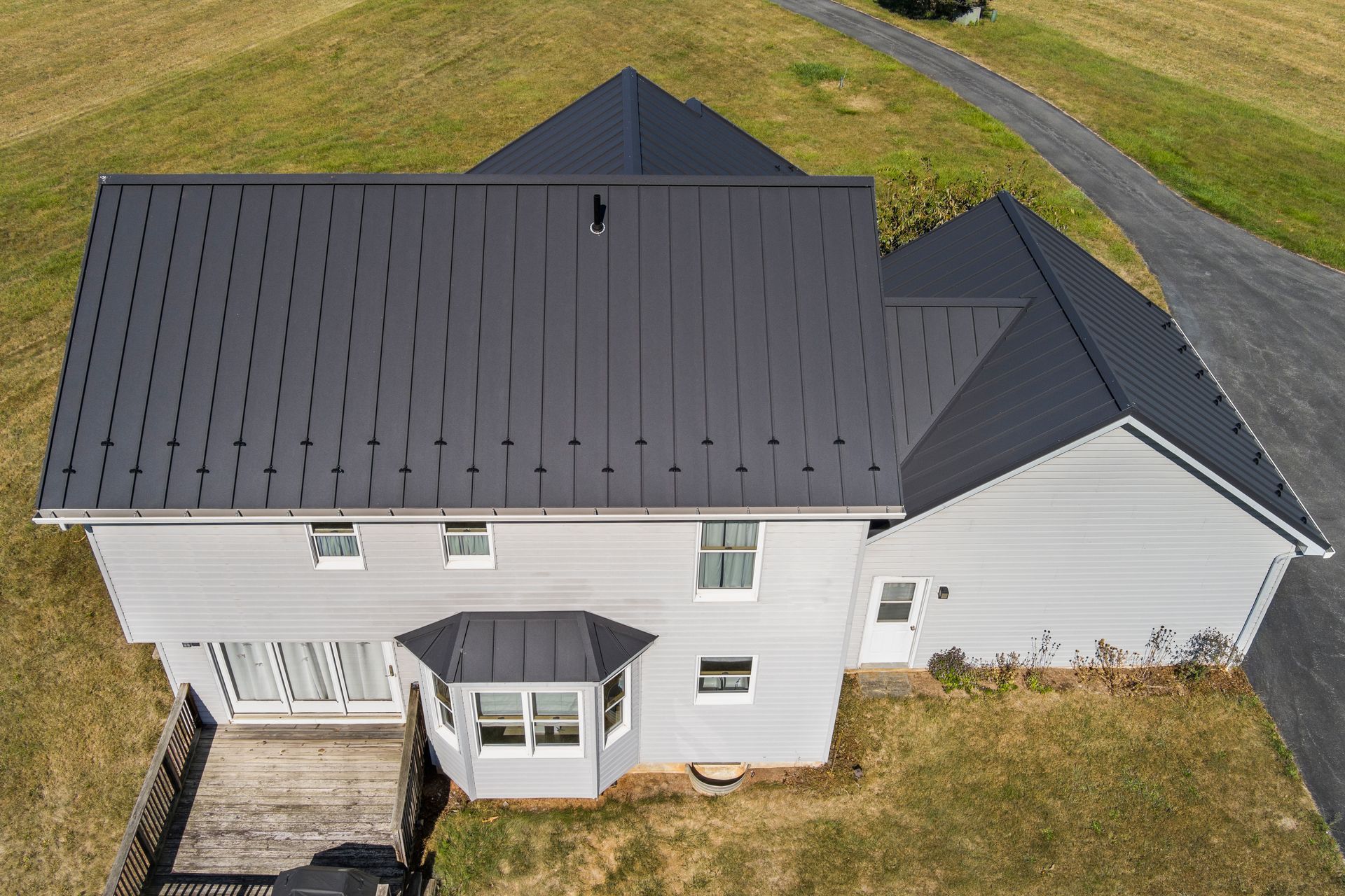 An aerial view of a two-story house with light gray siding, a dark metal roof, and a wooden deck, set in a grassy yard.