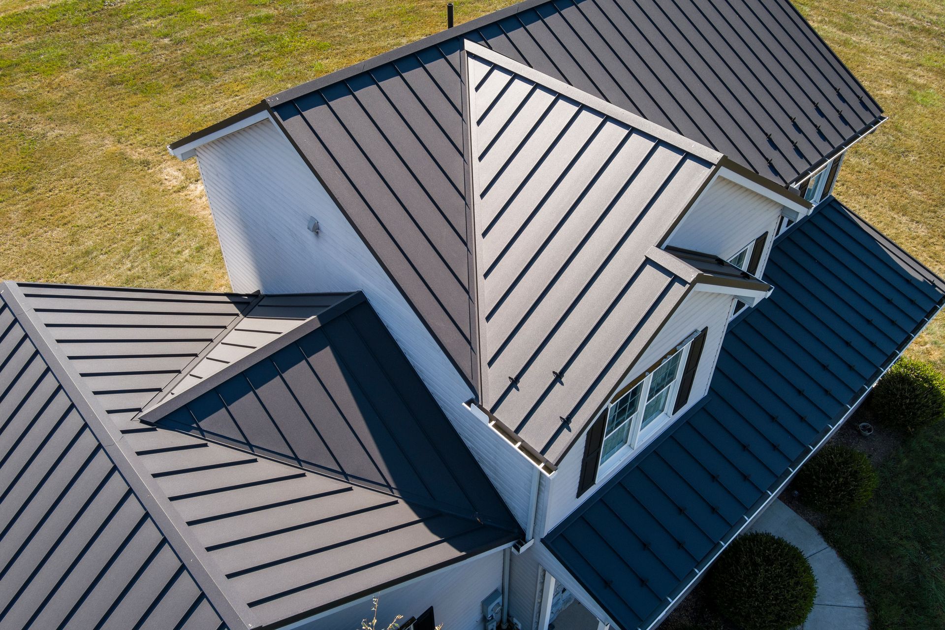 An aerial view of a white house with a dark metal roof, featuring multiple gables and a surrounding green lawn.