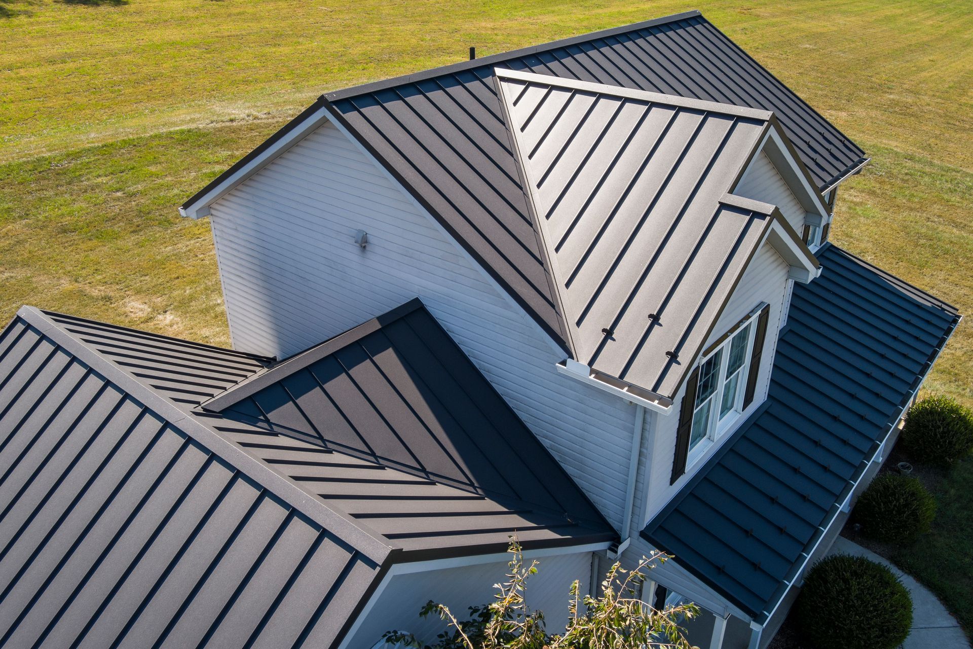 An aerial view of a white brick house with a dark, standing seam metal roof surrounded by green grass.