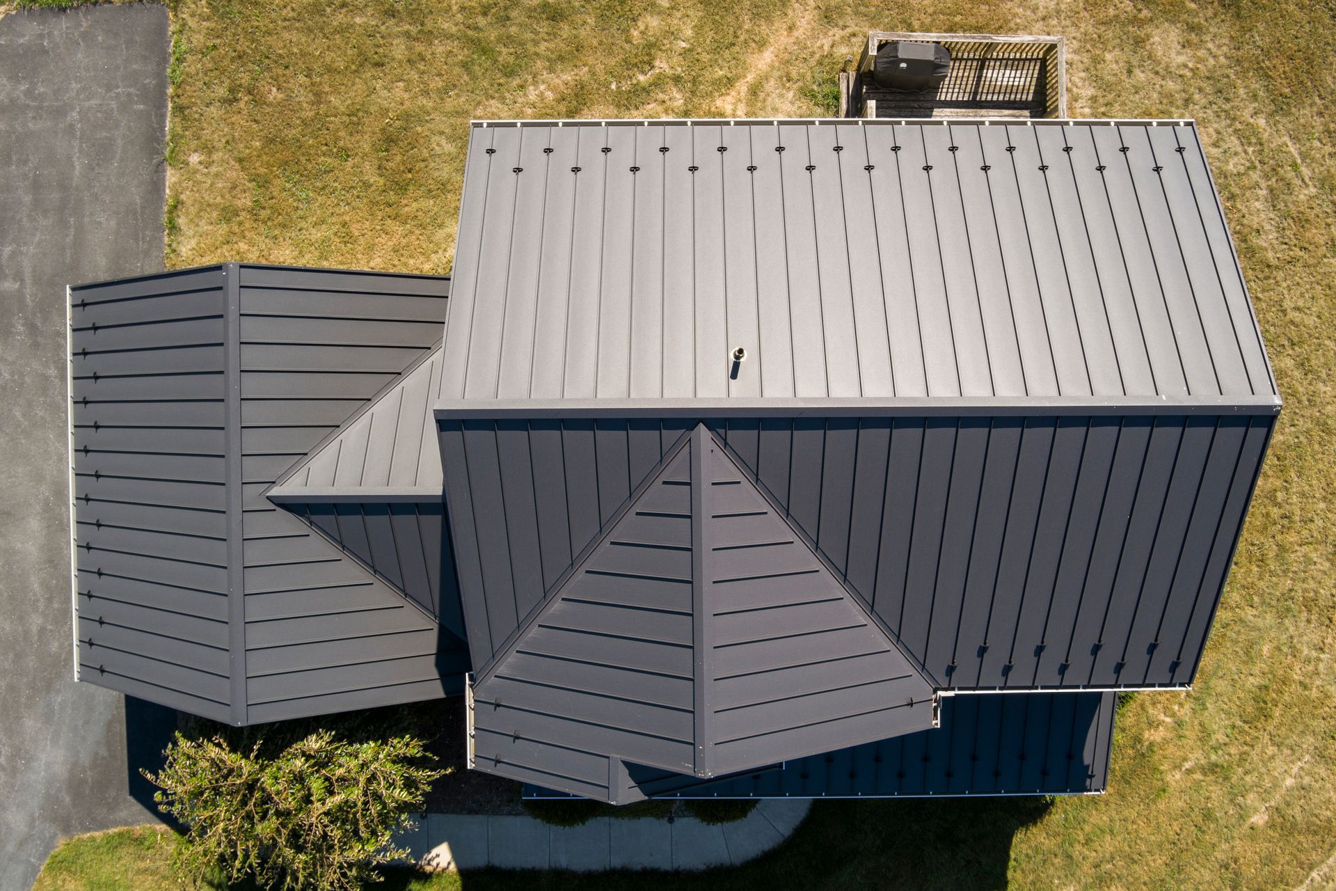 An aerial view shows a dark gray metal roof with several gables, surrounded by a grassy lawn and a paved driveway.