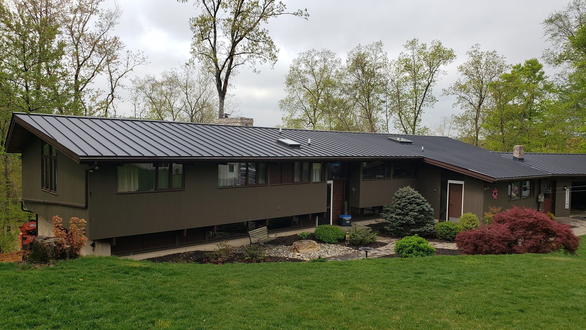 Brown, one-story house with a dark metal roof, built on a slight slope, surrounded by trees and a lawn.