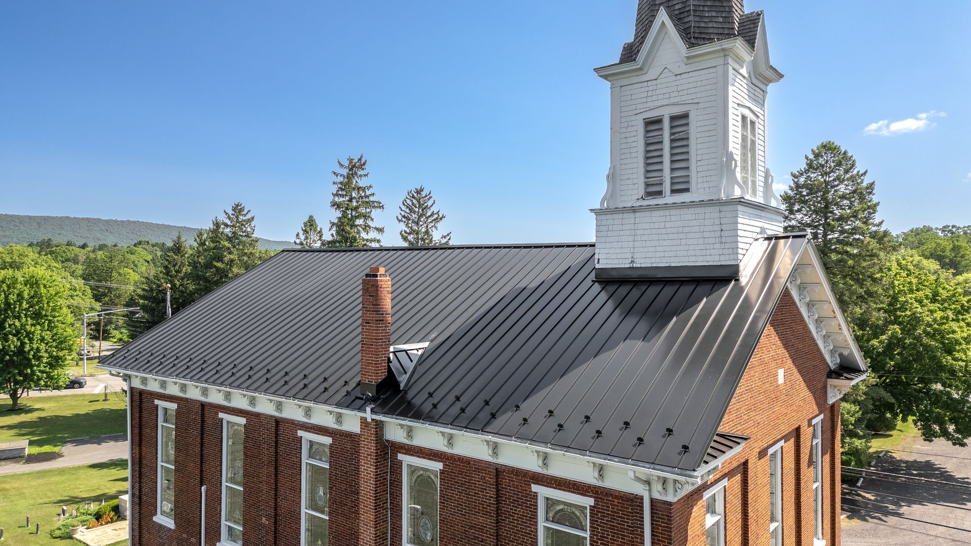 Brick church with a black metal roof and white steeple under a blue sky.