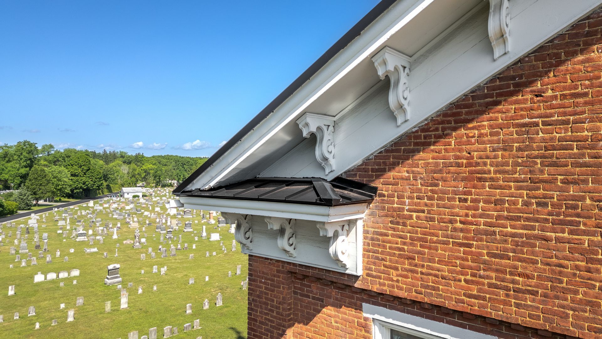Red brick building with white trim and decorative brackets, overlooking a cemetery on a sunny day.