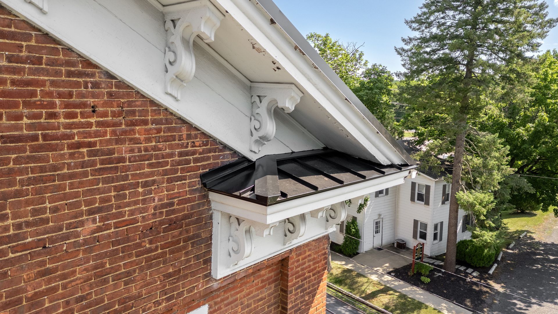 Brick building with white trim and black roof overhang; trees and house in background.
