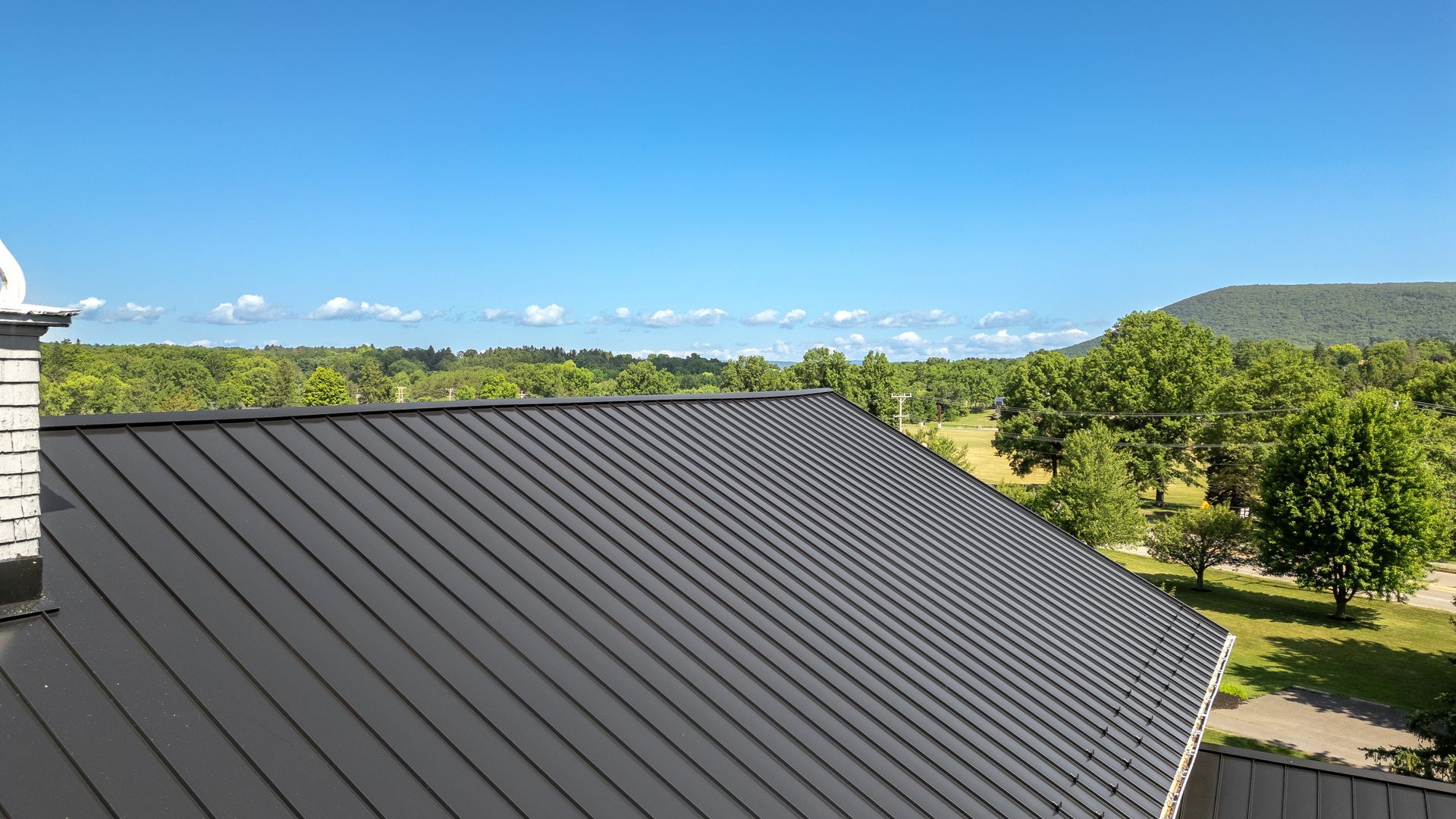 Dark gray metal roof with a scenic view of green trees, fields, and mountains under a blue sky.