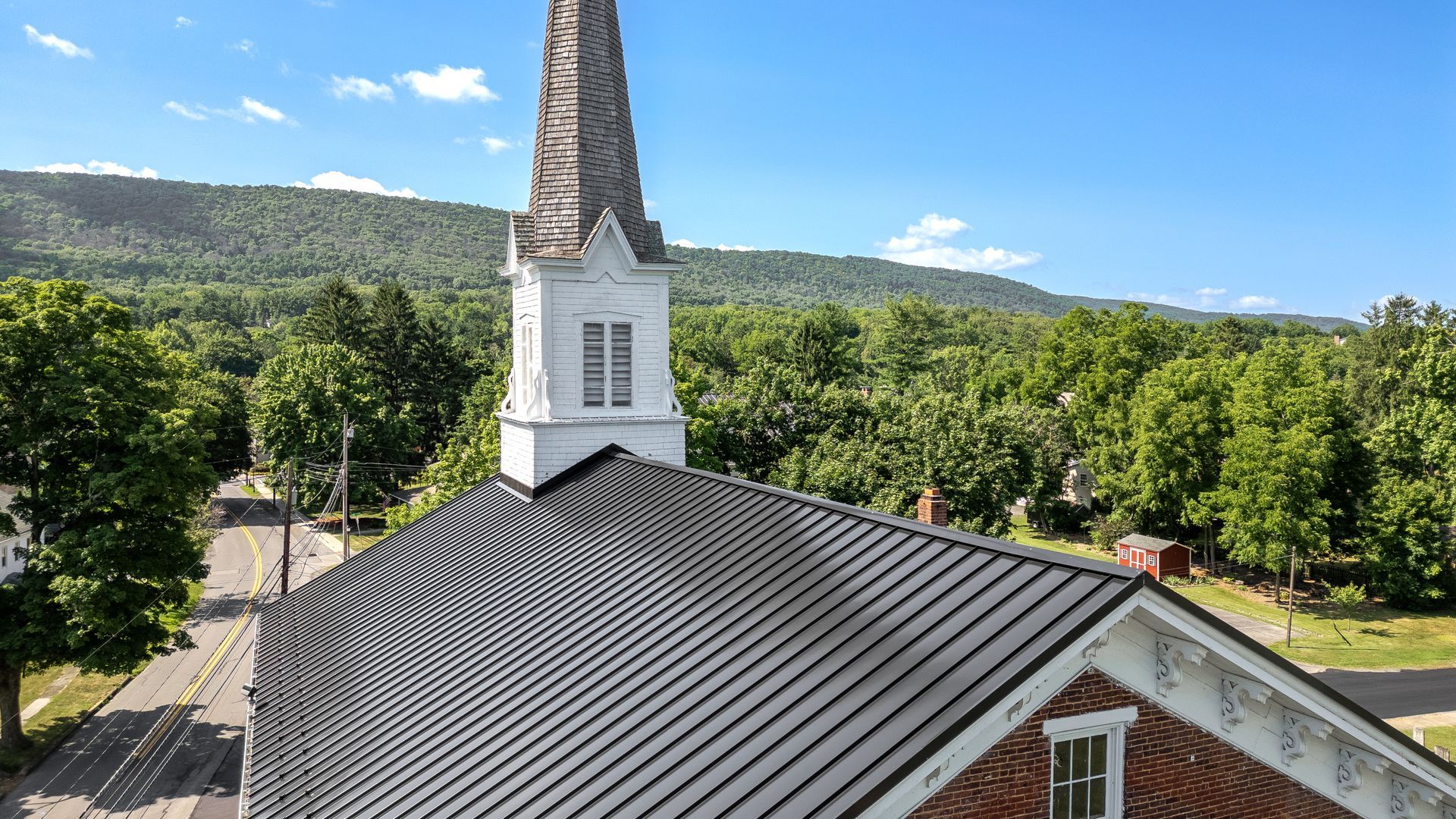 Church with a tall steeple, metal roof, and brick building against a backdrop of green trees and mountains.