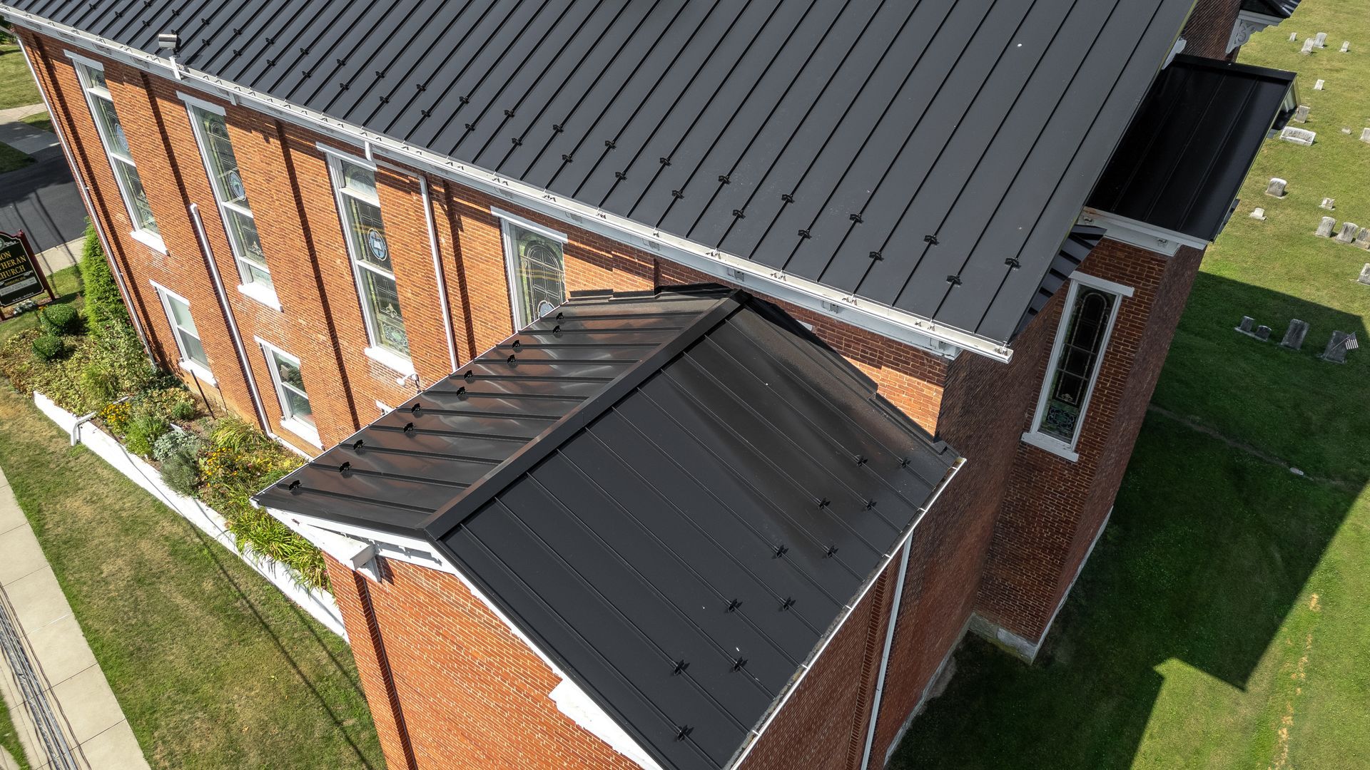 Black metal roof on a brick building with a grassy lawn and a cemetery in the background.