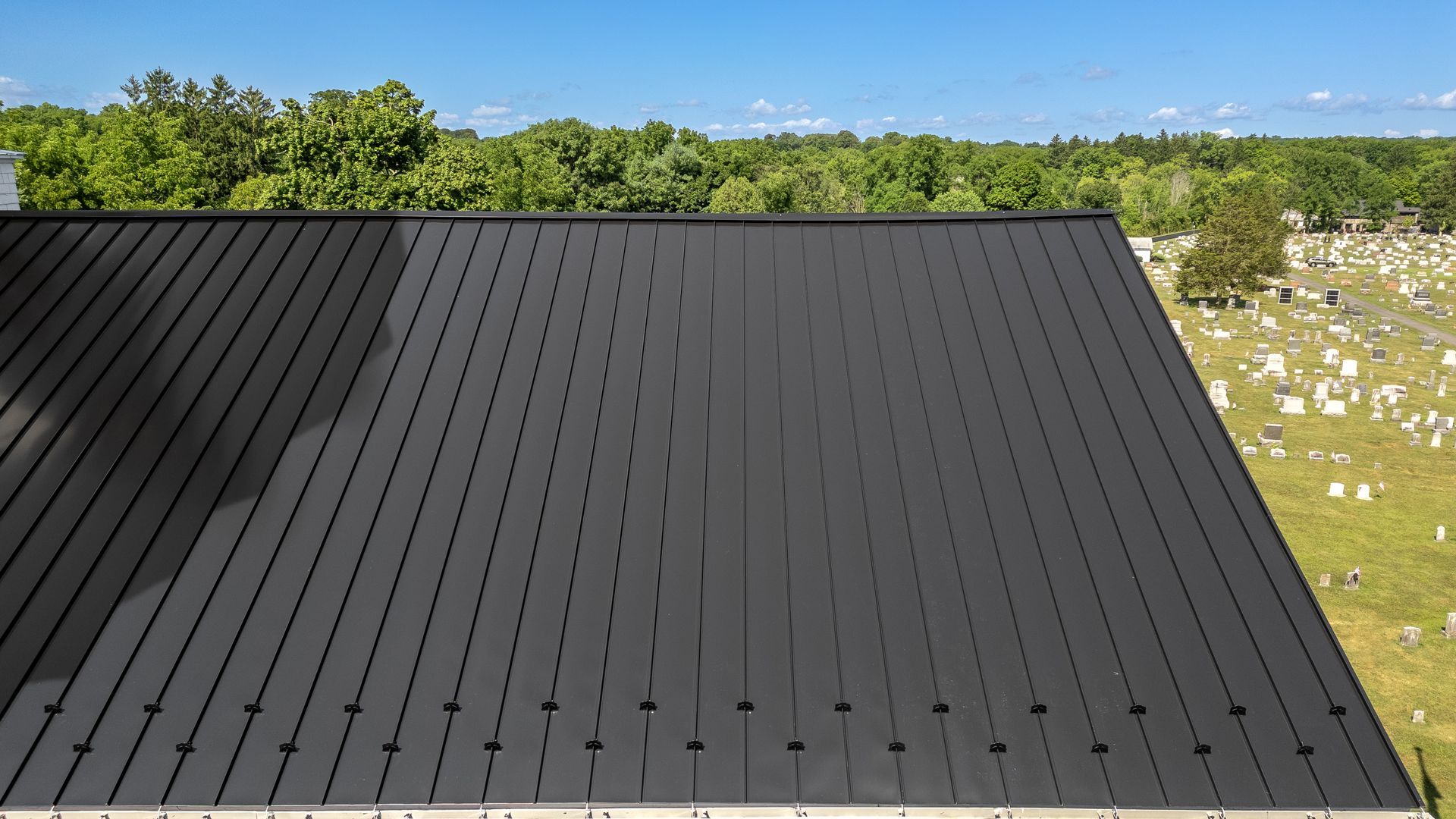 Black metal roof with a graveyard and trees in the background under a blue sky.