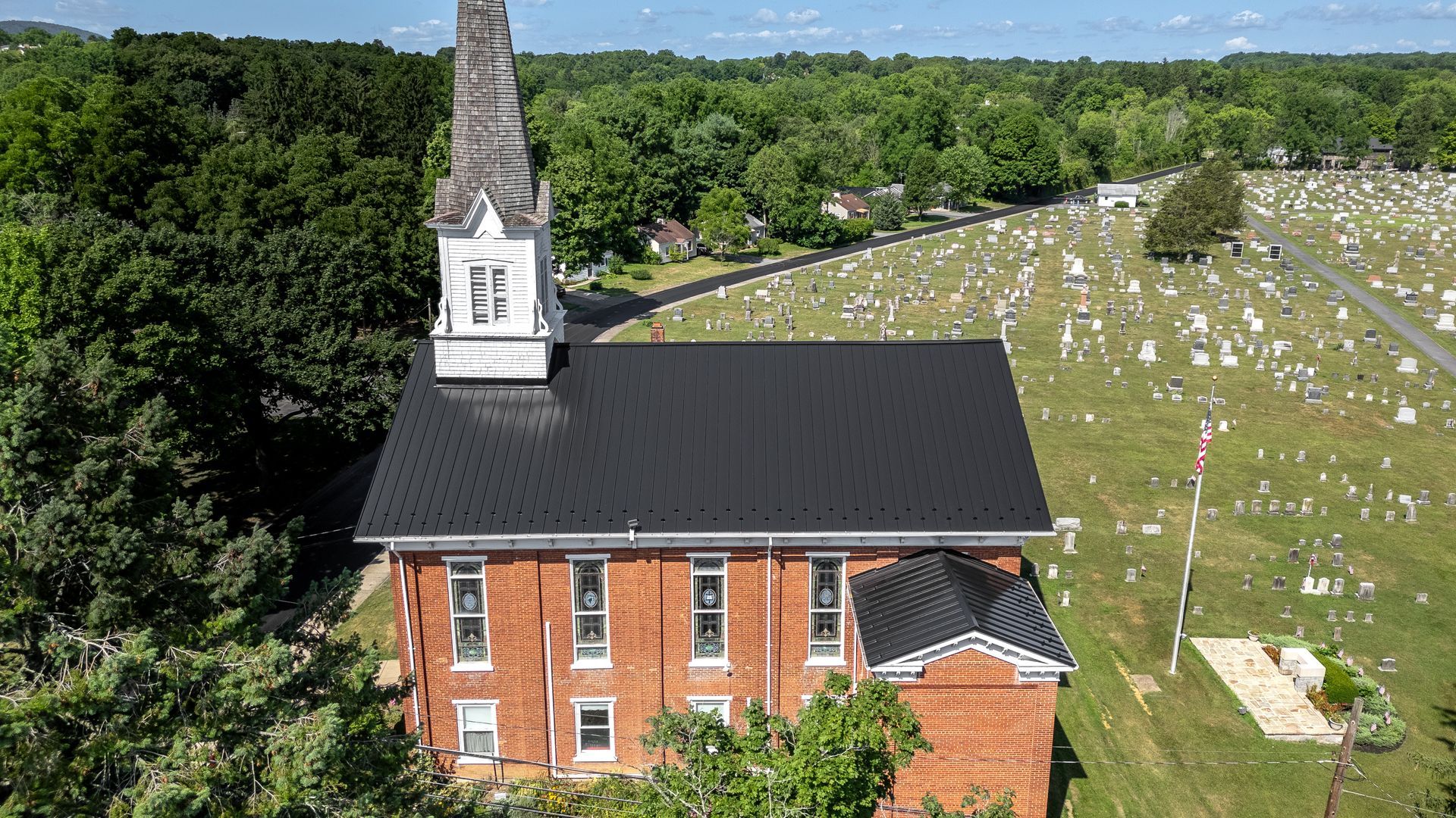 Brick church with tall steeple next to a cemetery, trees in the background.