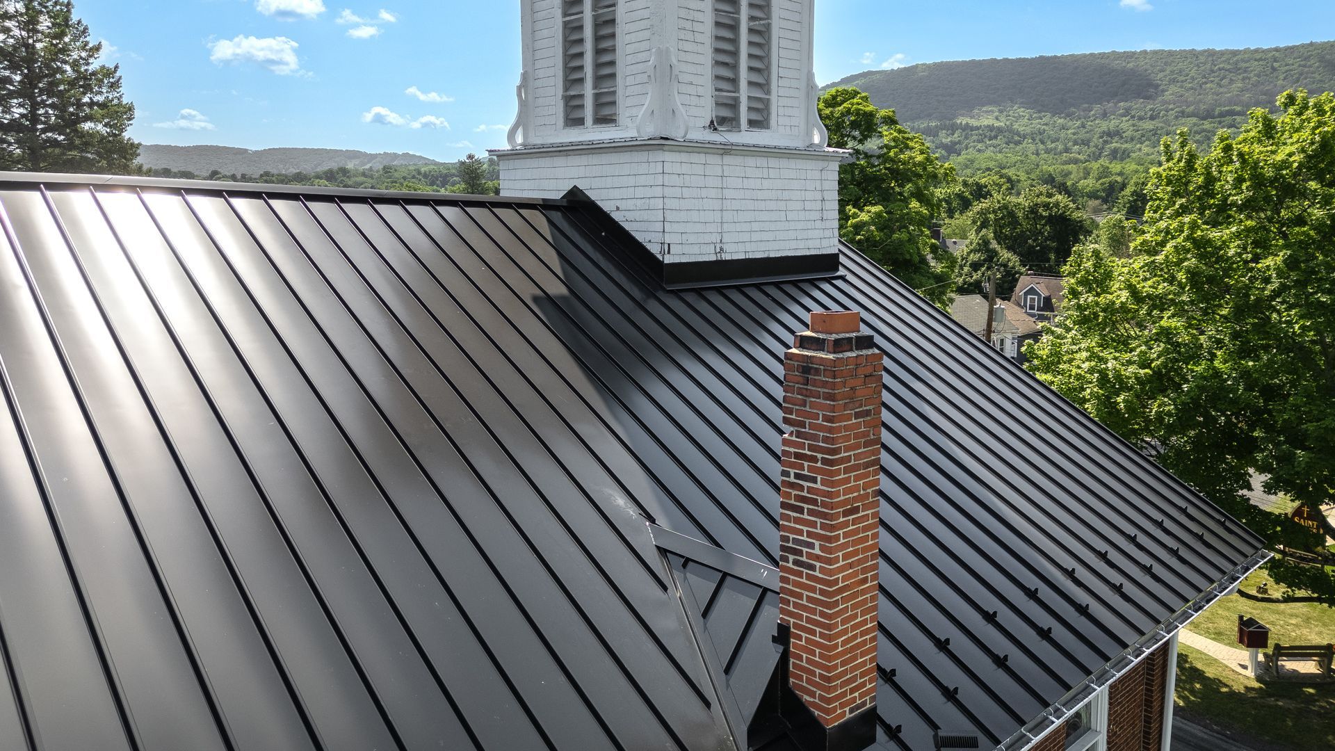 Black metal roof on a building with a brick chimney and white steeple, with green trees and mountains in the background.