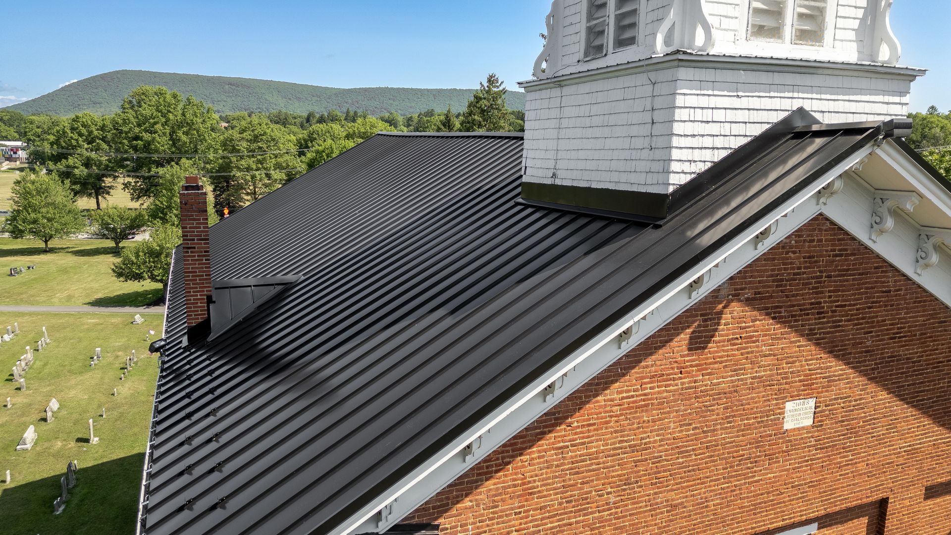 Brick building with dark metal roof; steeple in background, green trees, and mountains.