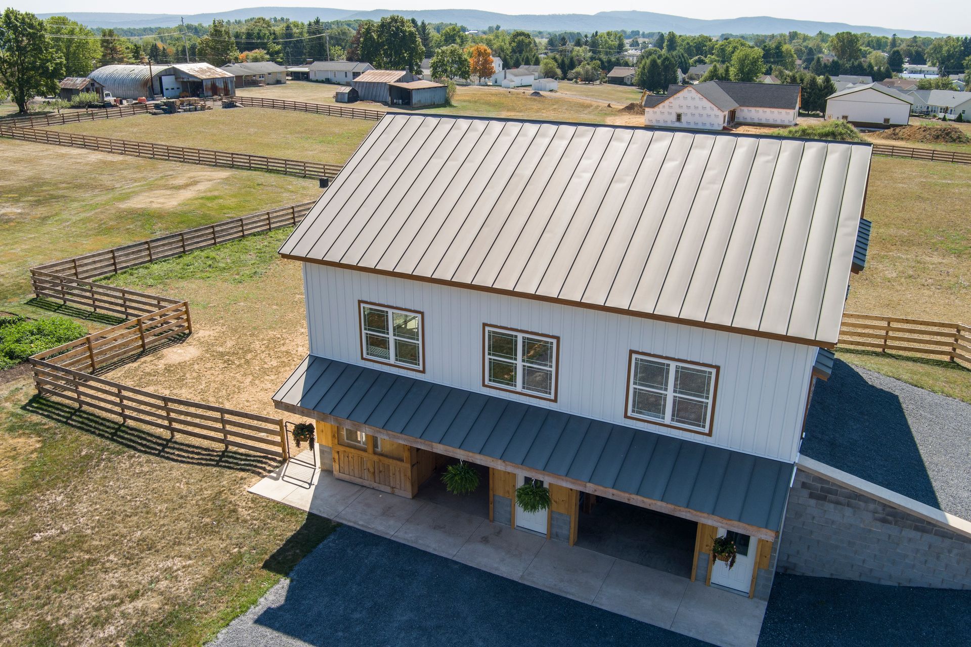 Aerial view of a white, two-story barn with a dark metal roof, set in a sunny, fenced rural landscape.