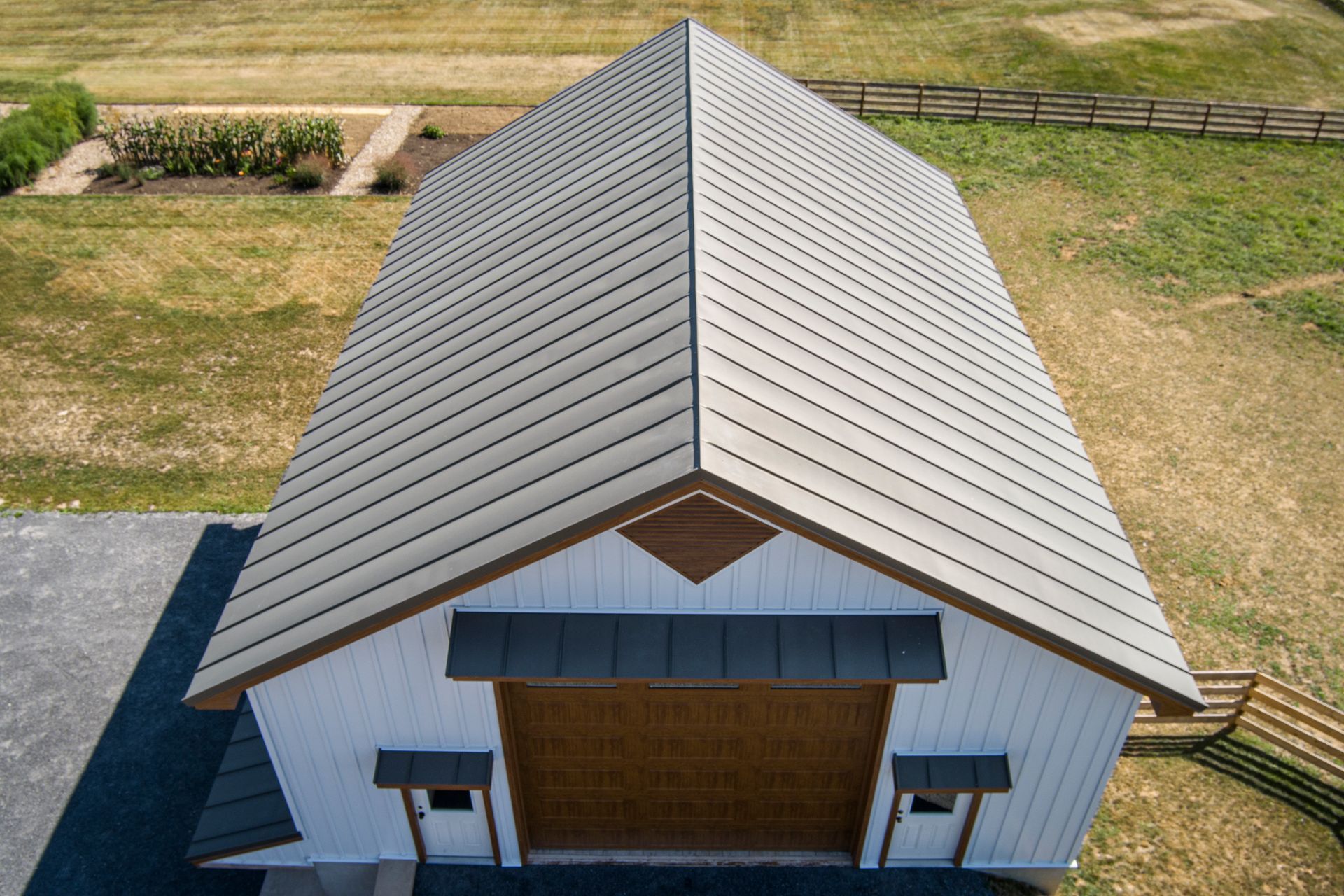 An aerial view of a white barn with a tan metal roof, a large wooden garage door, and a grassy, fenced-in yard.
