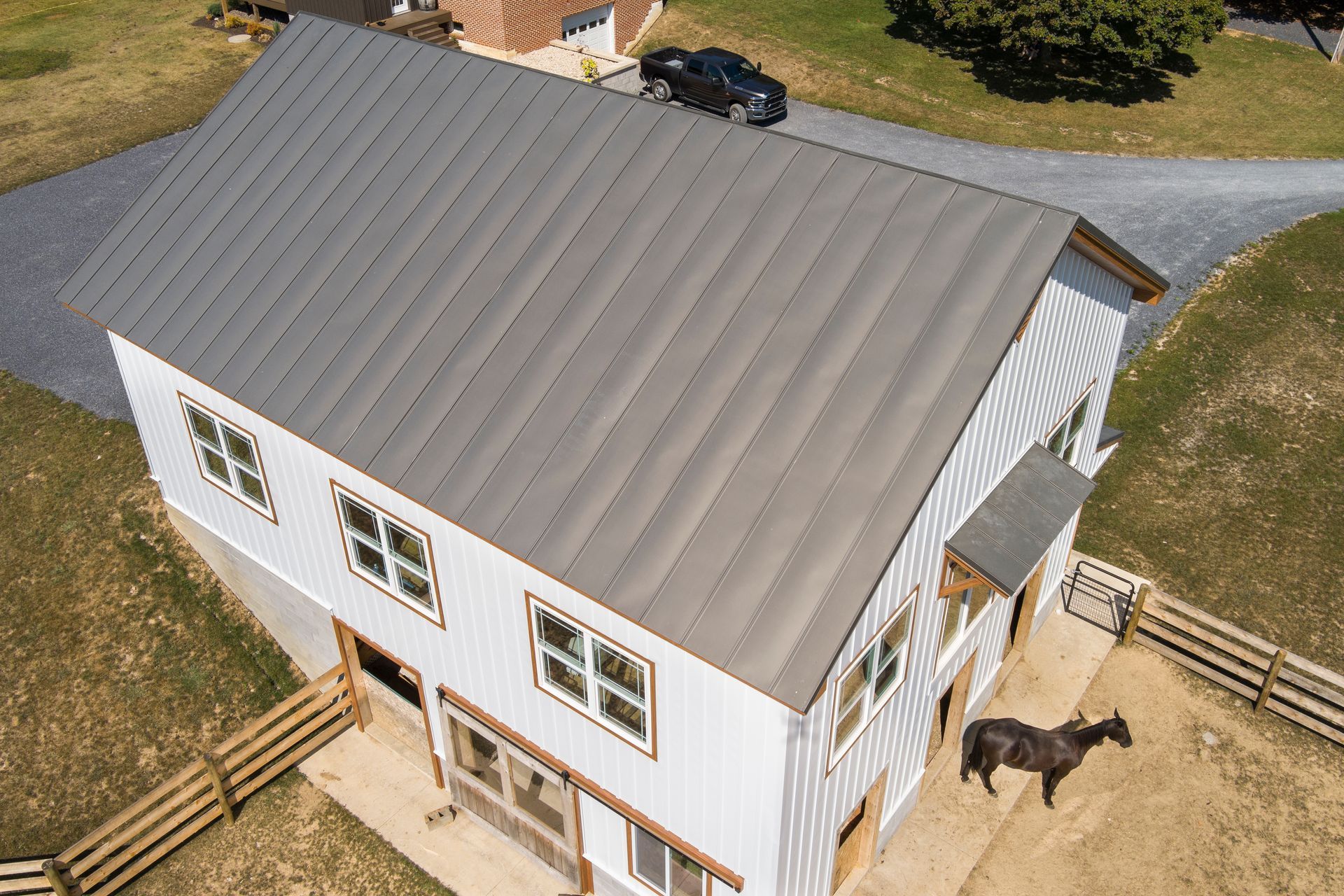Aerial view of a white barn with a dark metal roof, located in a fenced grassy lot with a horse and a parked truck.