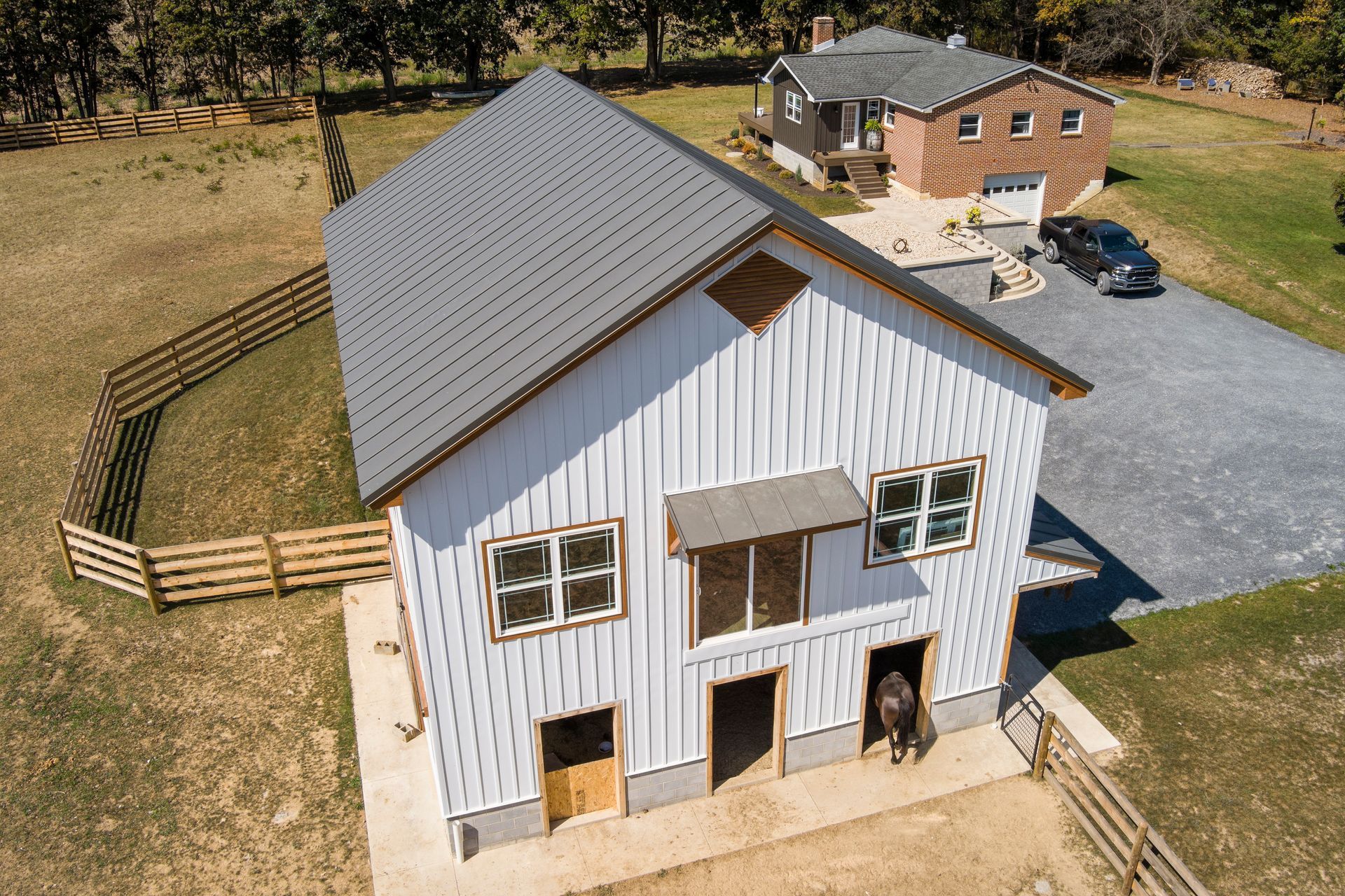 An aerial view shows a white barn with a dark gray metal roof next to a brick house on a rural property with fenced pastures.