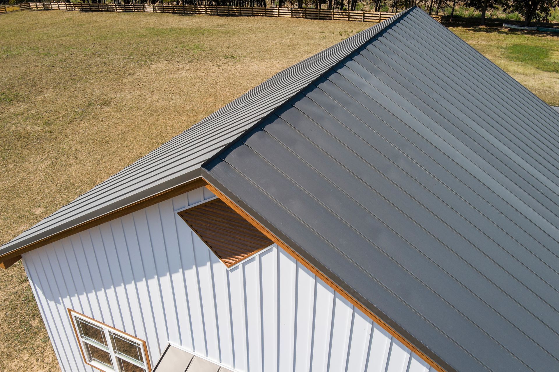 An elevated view of a white building with vertical siding and a dark gray metal roof, set against a dry grassy field.