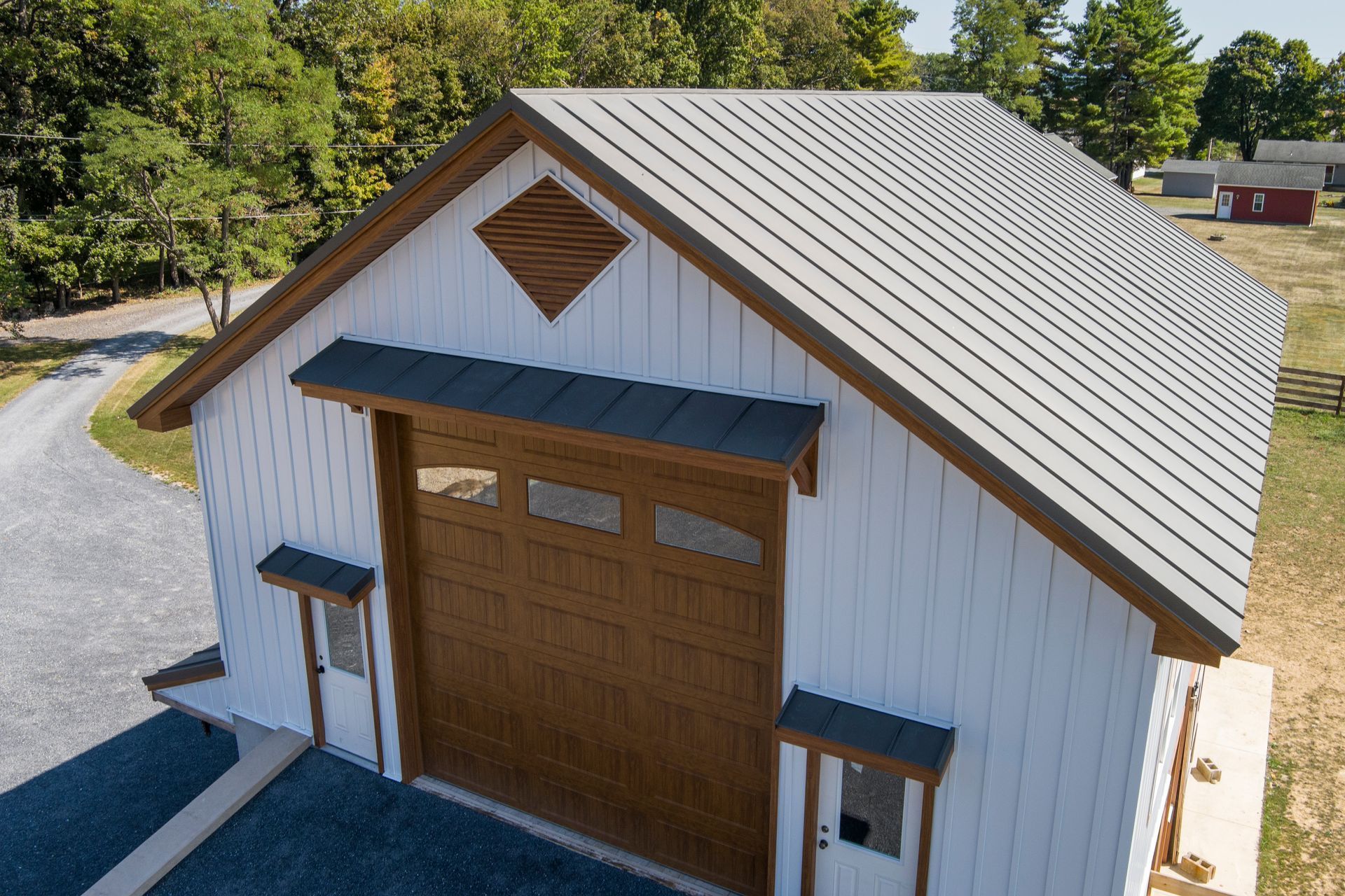 A white, metal gable-roofed garage with brown trim, a large brown door, and two small entrance doors on a gravel driveway.