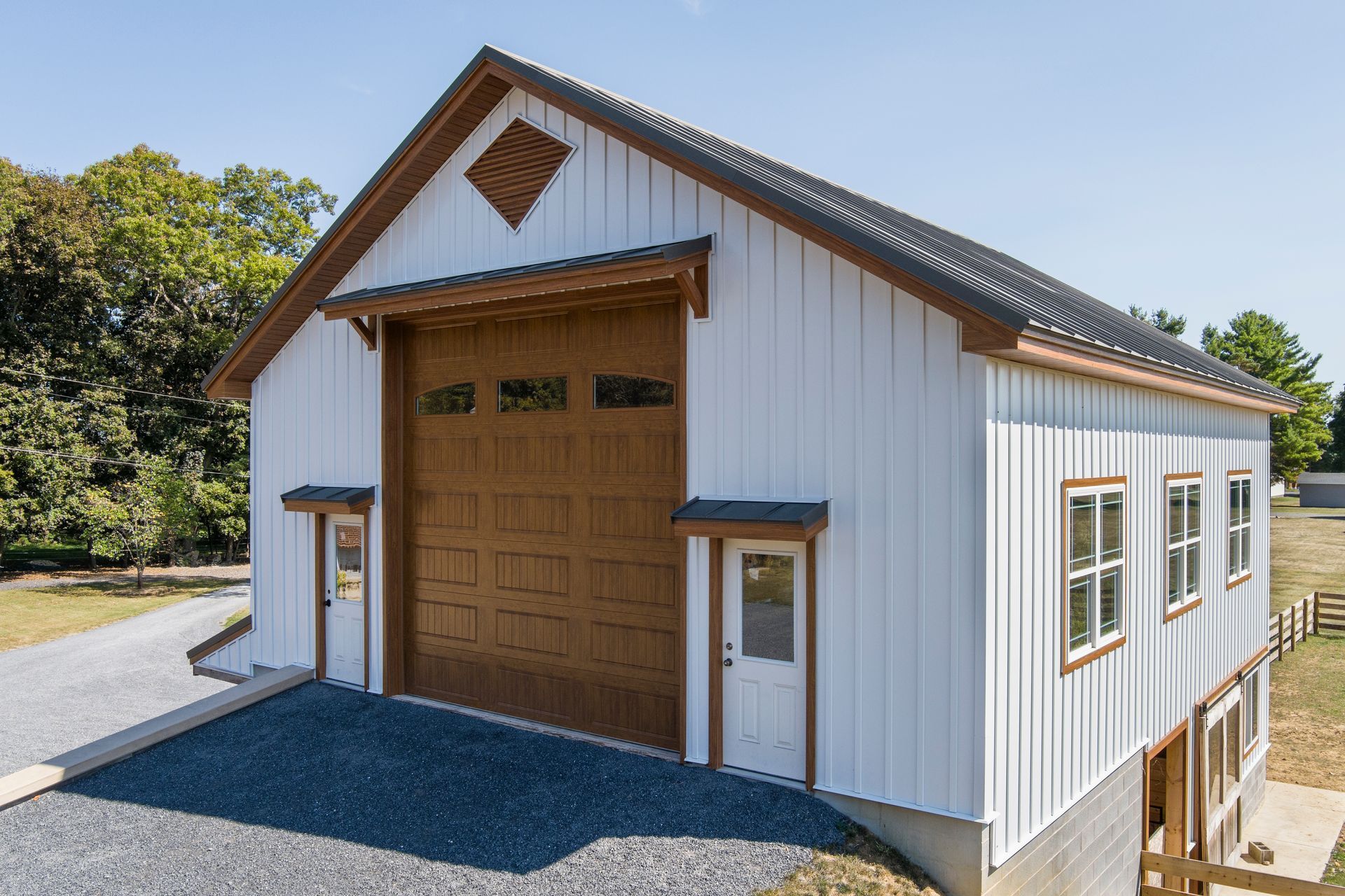 A two-story white barn-style building with a large brown garage door, wooden trim, and a metal roof on a gravel lot.