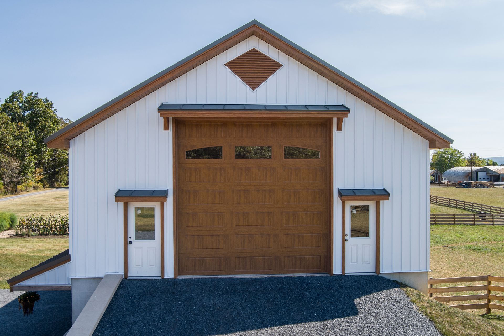 A white barn with a large brown garage door, two white pedestrian doors, and a decorative diamond-shaped gable vent.