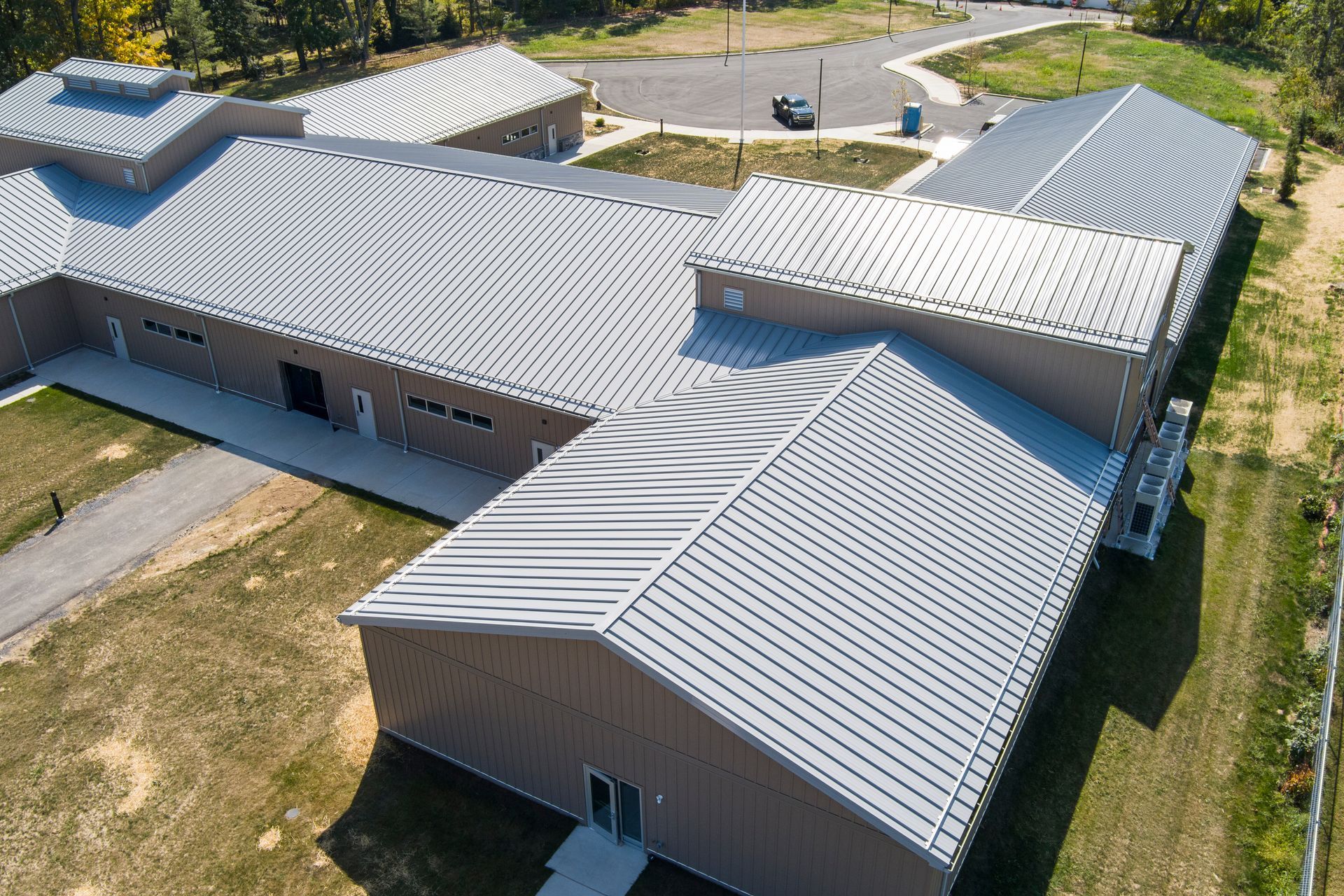 An aerial view of a tan, L-shaped building with a corrugated metal roof, surrounded by lawns and a gravel driveway.