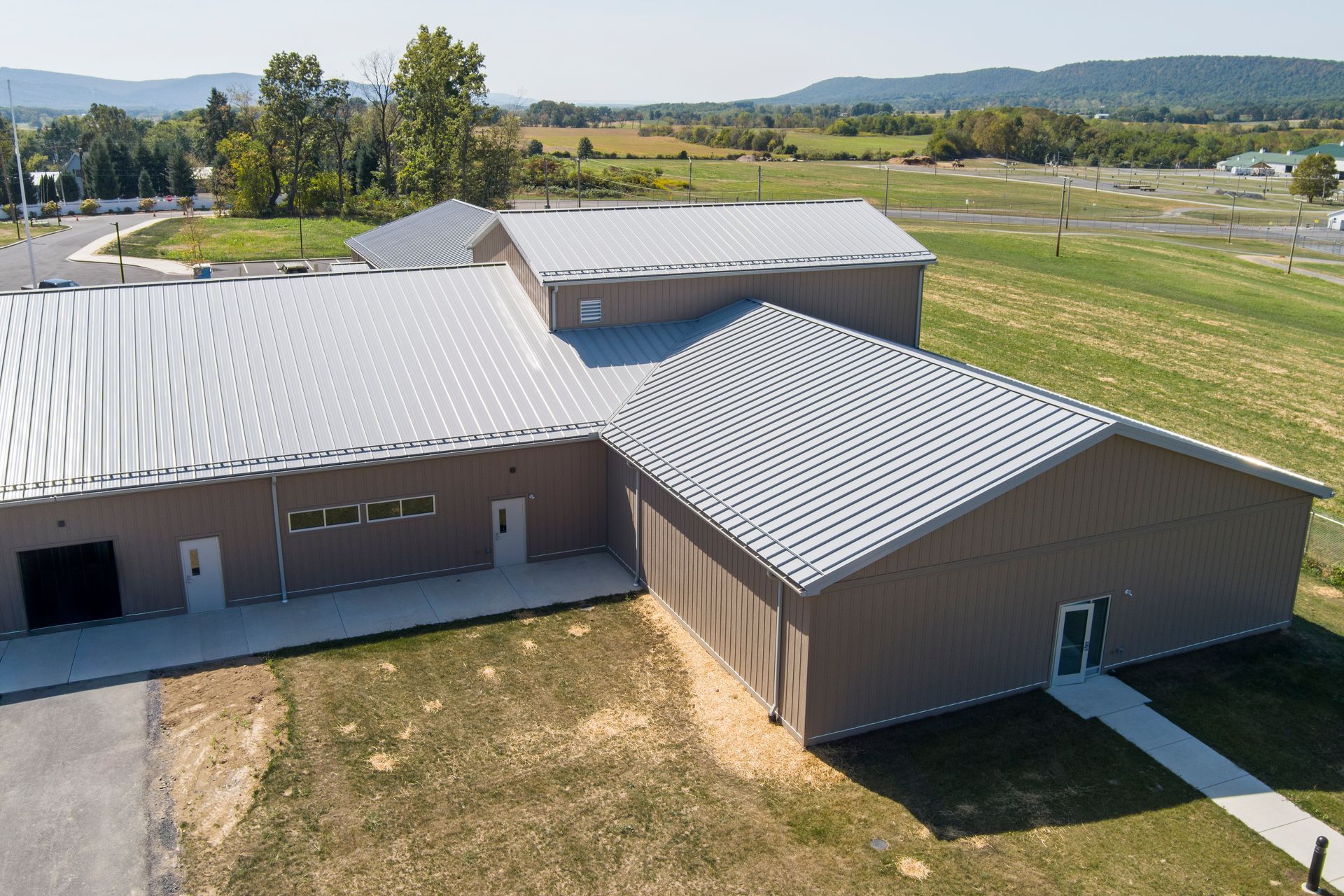 An aerial view of a tan, single-story commercial building with a grey metal roof, set against a grassy field and hills.