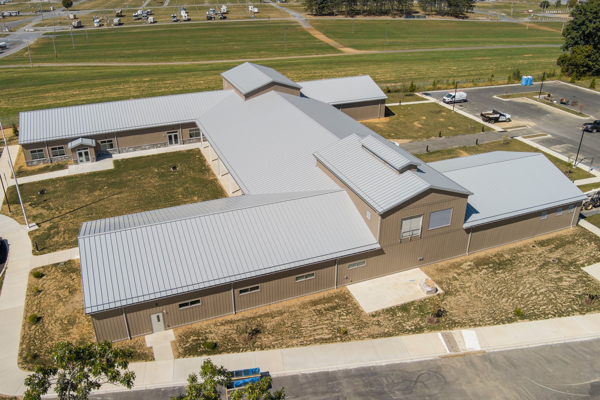 An aerial view of a tan, U-shaped single-story building with a light-colored metal roof surrounded by lawn and parking.