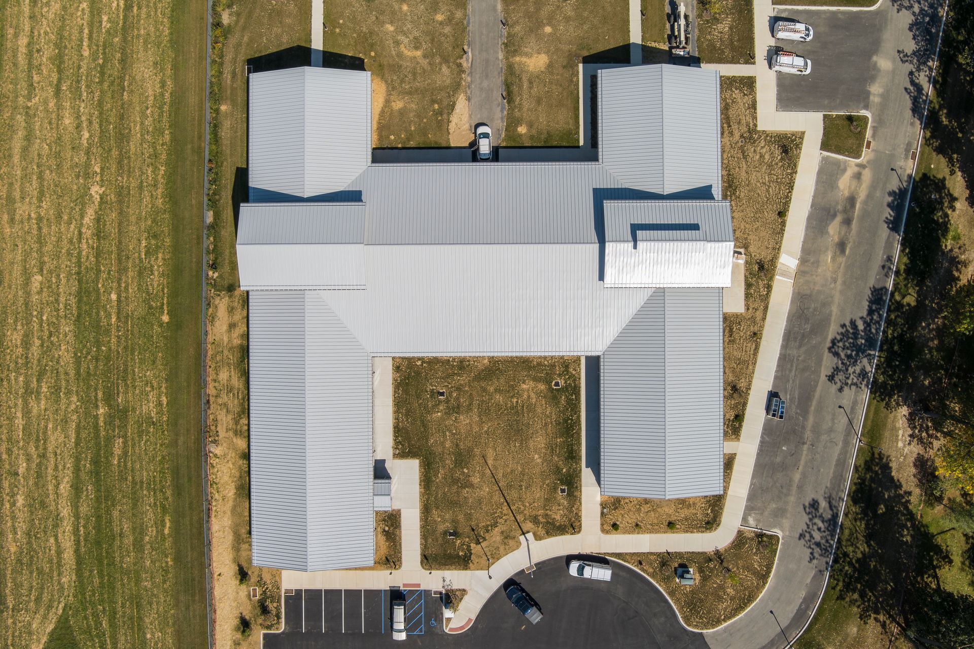 Top-down aerial view of a building with a light-colored metal roof, surrounded by parking and grassy areas.