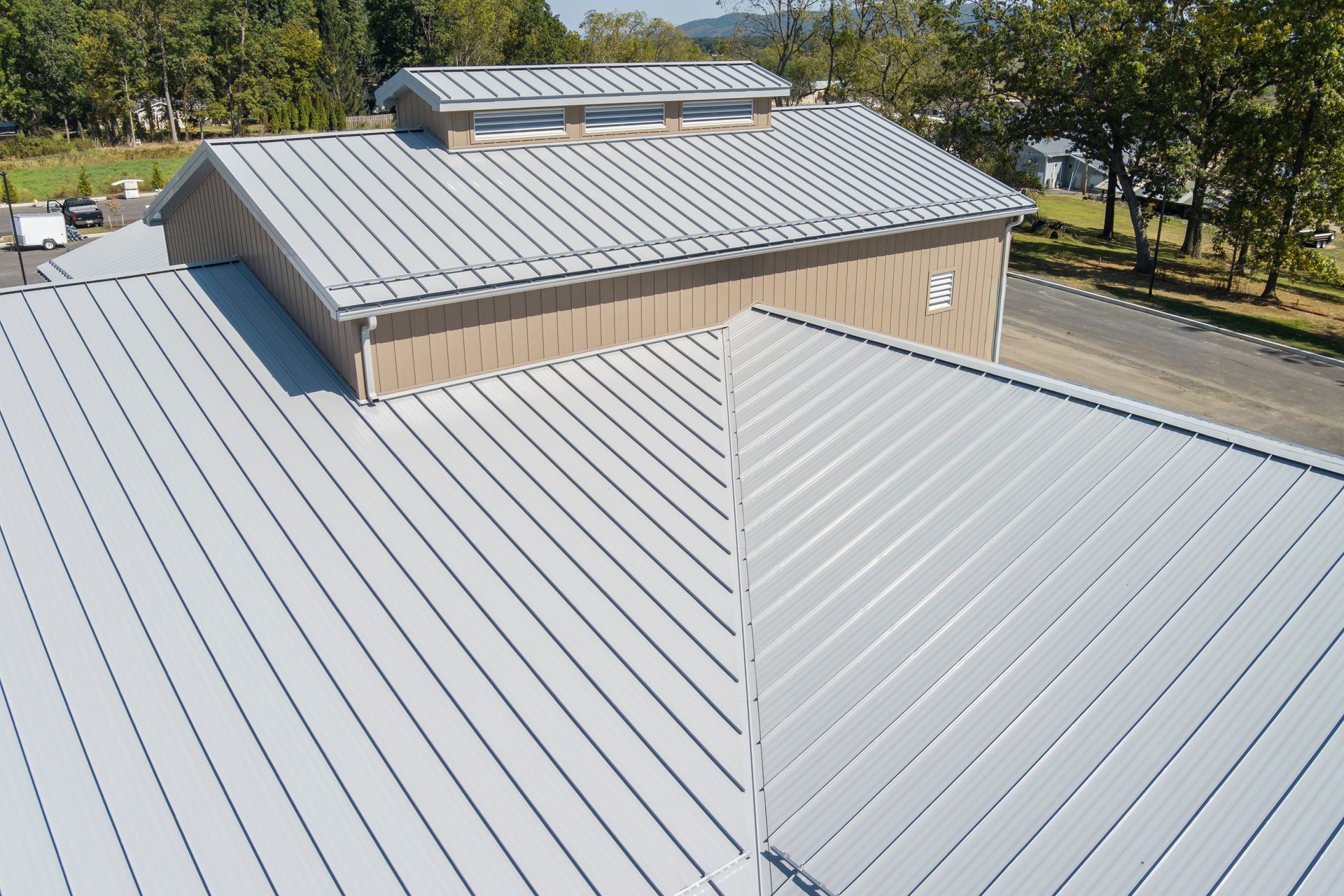 An elevated view of a light gray metal roof with a central clerestory window section, surrounded by trees.
