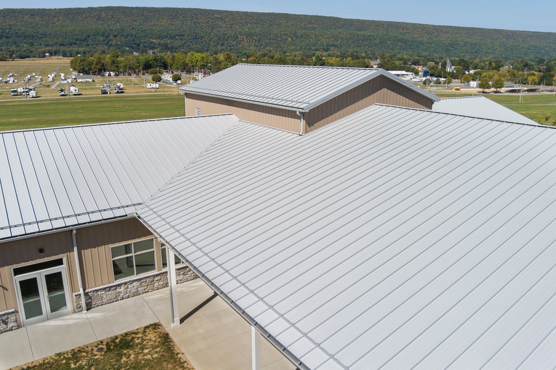 An elevated, angled view of a modern building with a light gray metal roof, set against a green, rolling landscape.