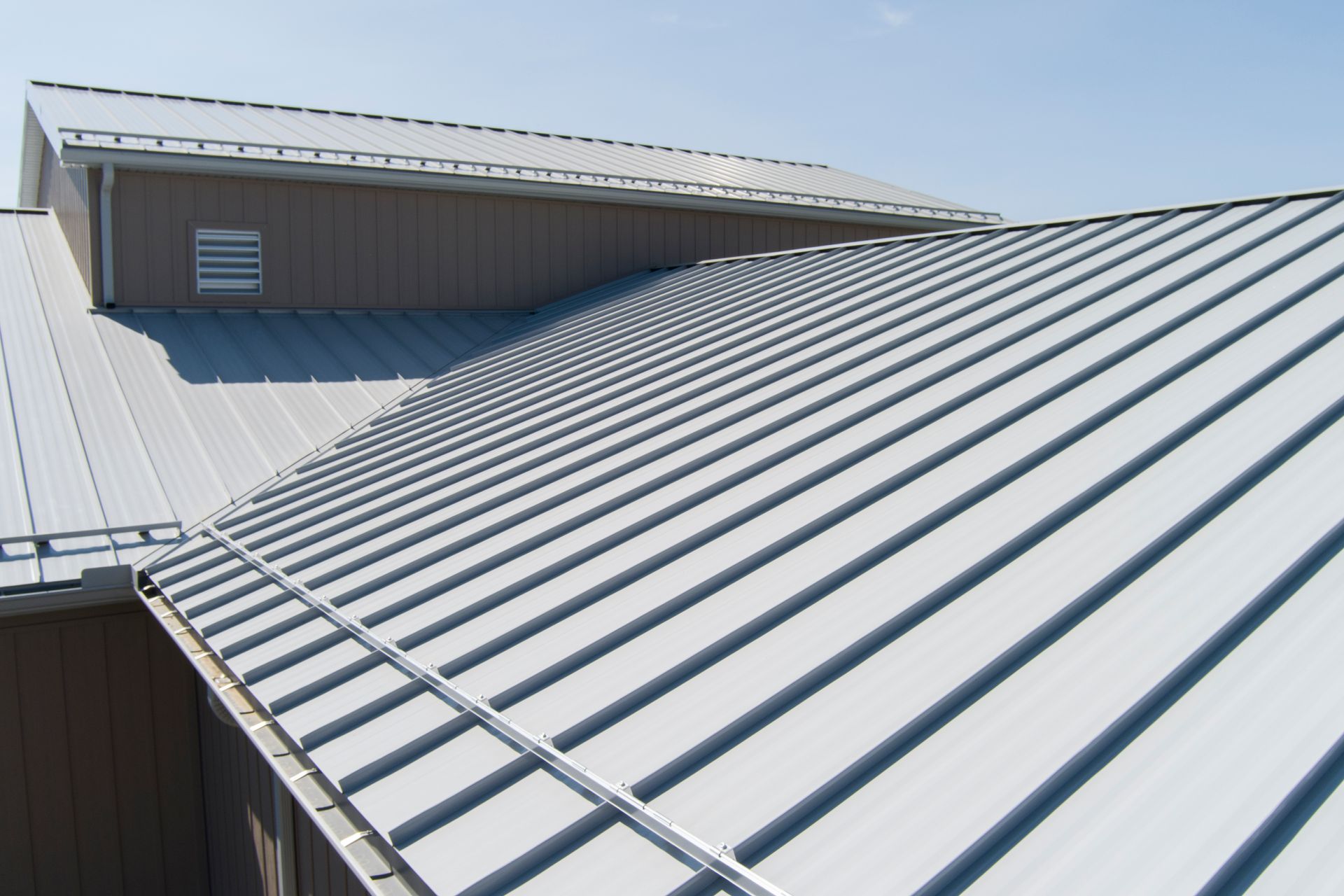 A close-up view of a metal roof with light gray vertical panels and a tan-colored wall section under a clear blue sky.