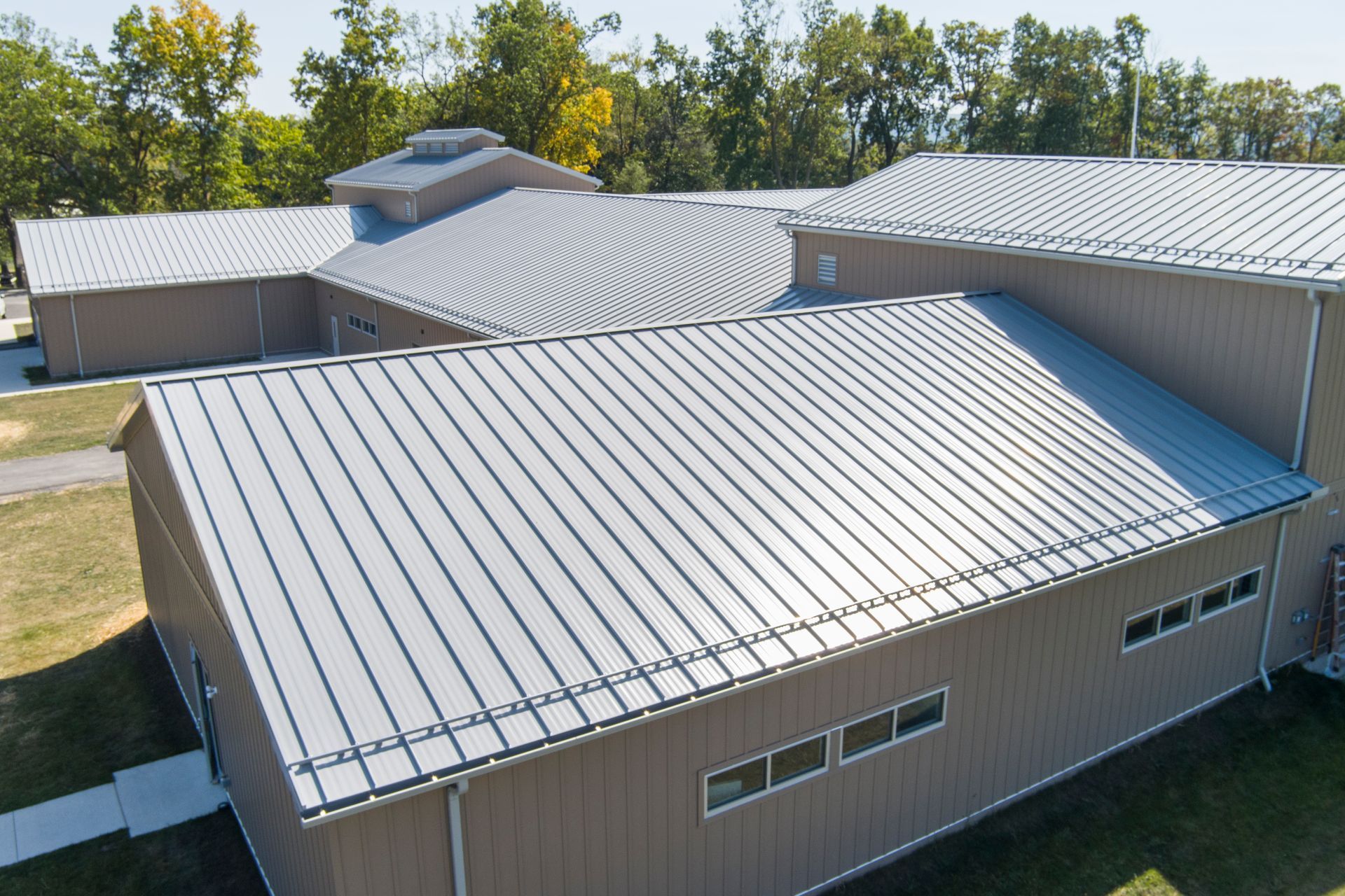 An aerial view of a tan, multi-section building featuring a light grey metal roof, set against a backdrop of trees.