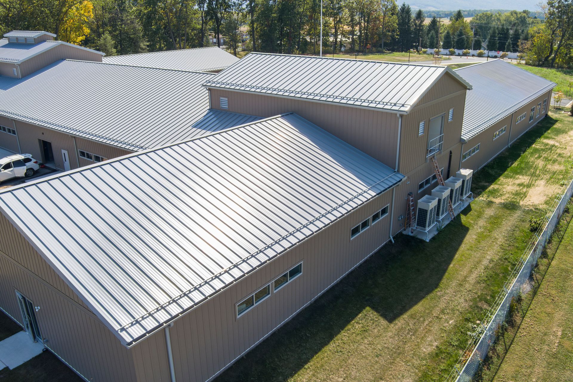 Aerial view of a long, tan industrial building with a silver, ribbed metal roof, surrounded by grass and trees.