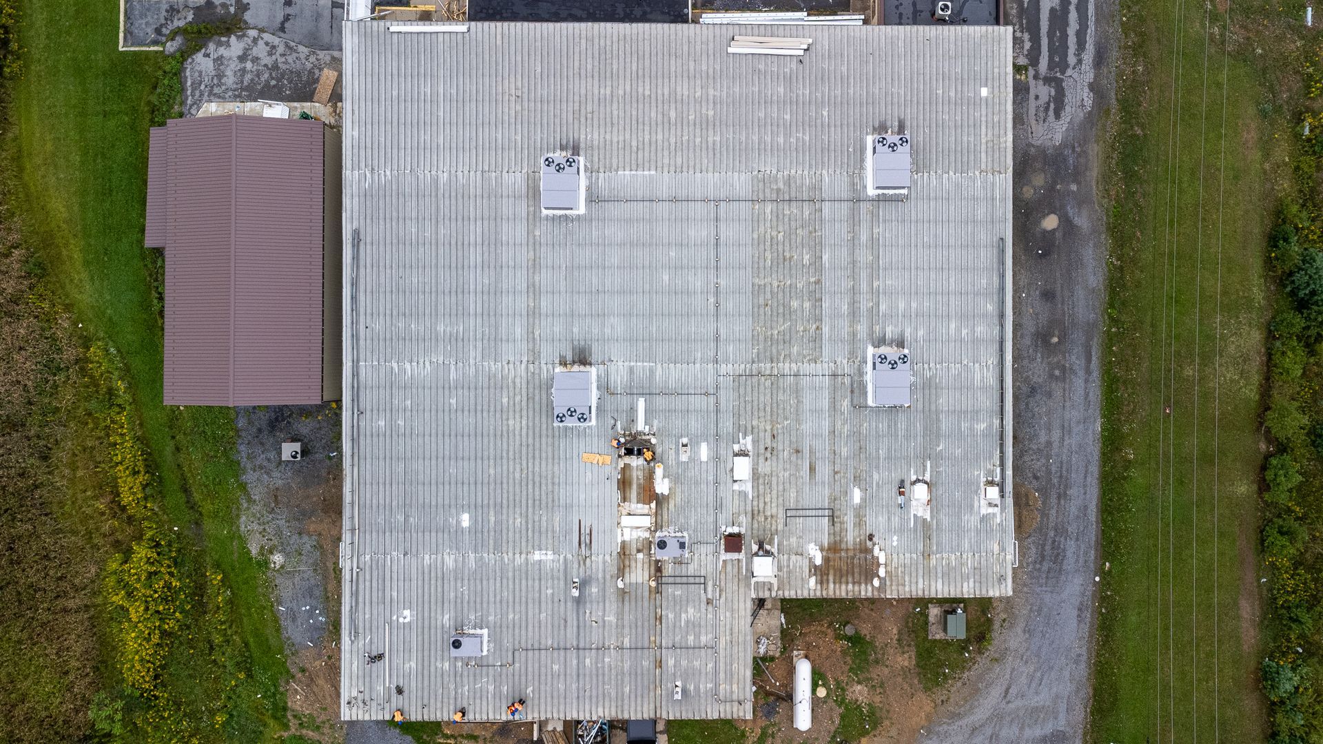 Aerial view of a large gray building with a damaged roof, surrounded by green grass and a brown building.