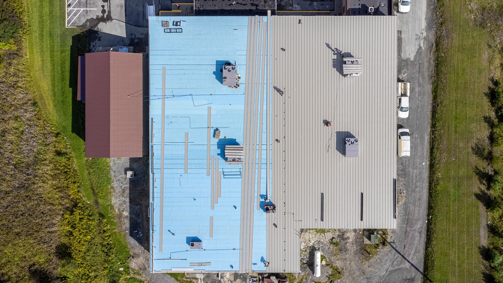 Overhead view of a building with a blue and silver metal roof, surrounded by green grass and a dirt path.