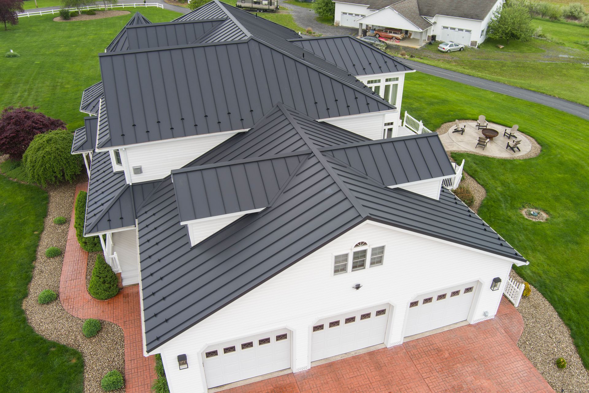 White house with dark gray metal roof, three-car garage, and red brick driveway, surrounded by green grass.