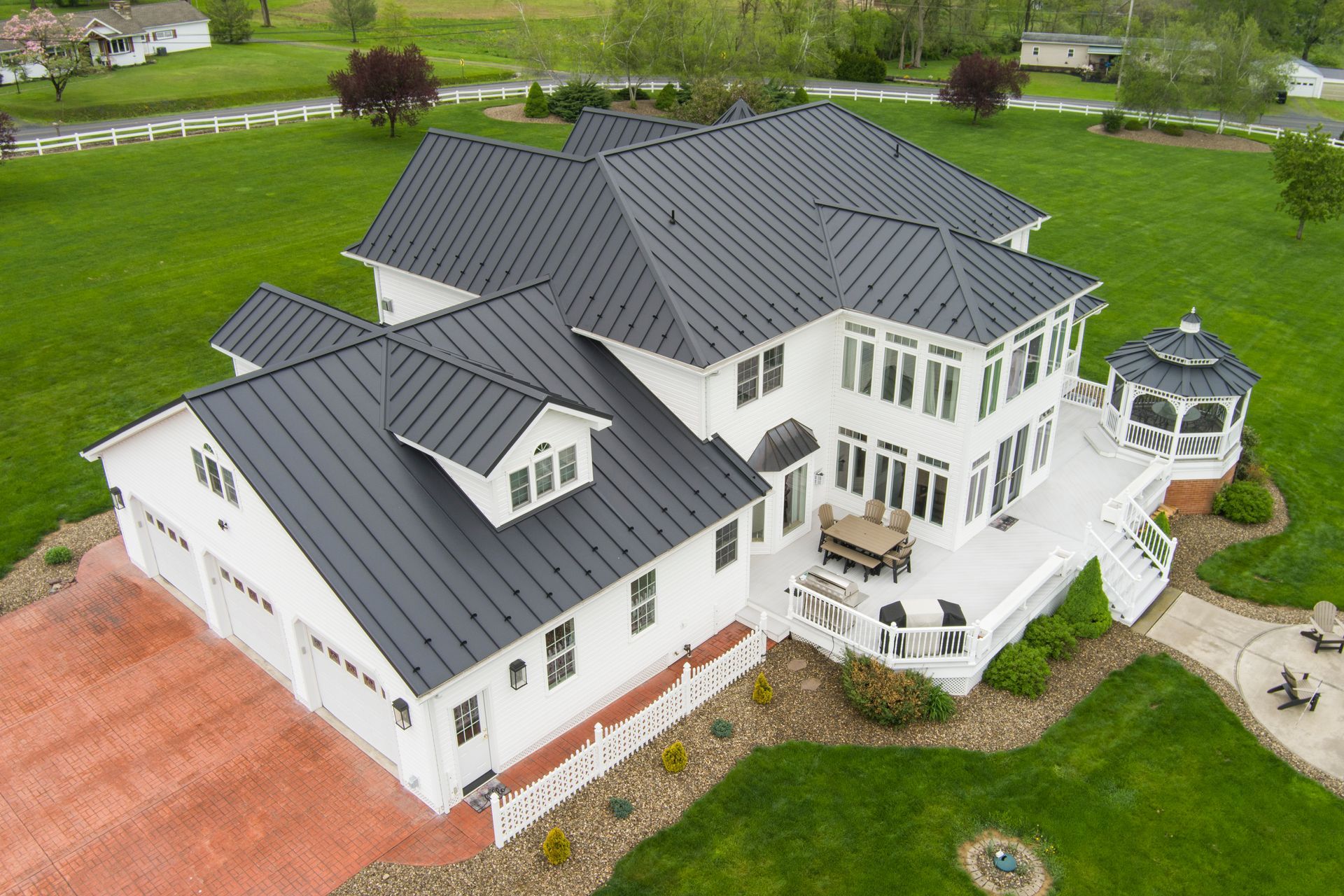 White house with black roof, red brick driveway, and gazebo on a large green lawn.
