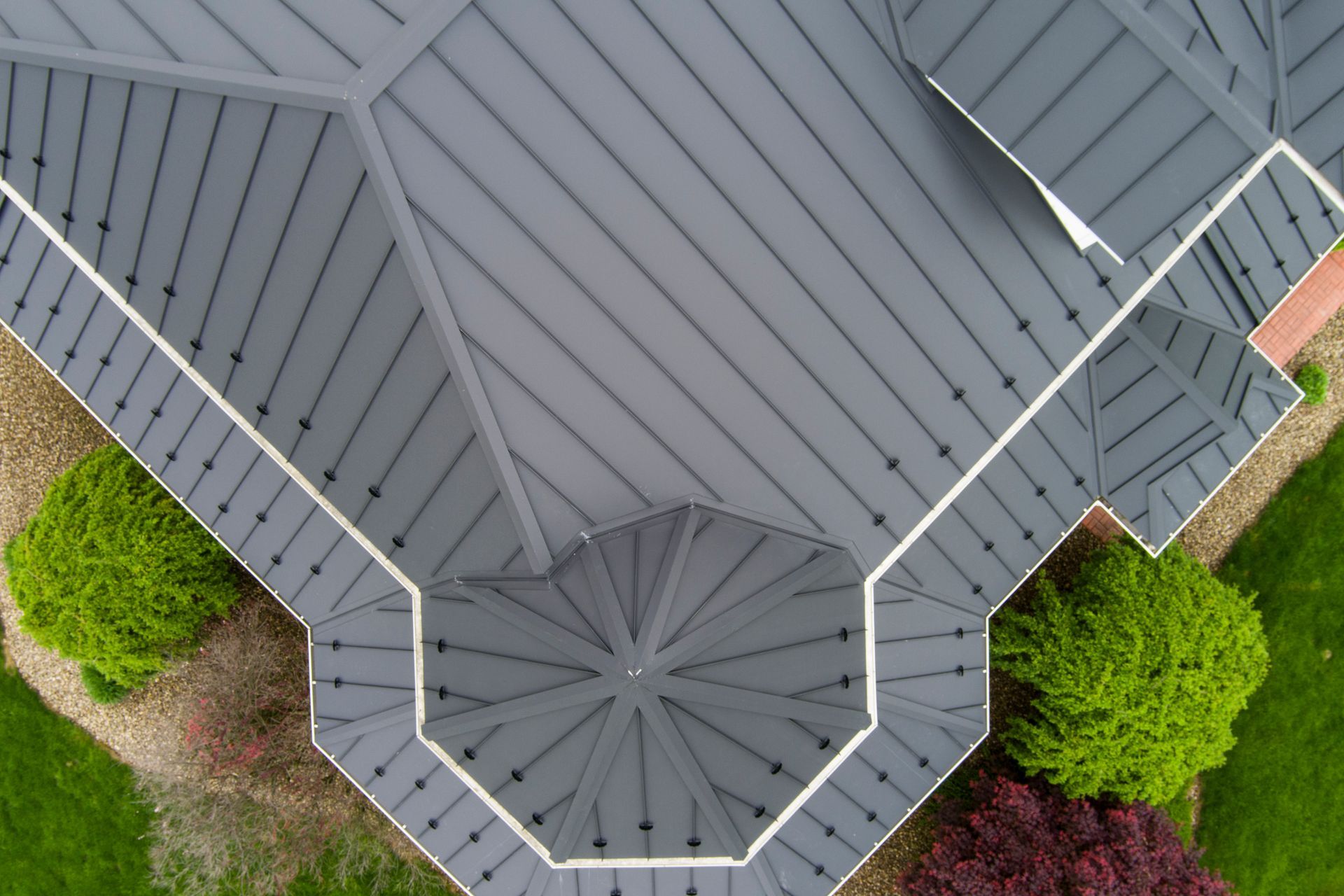 Overhead view of a gray metal roof with a circular portion and surrounding landscaping with green bushes.