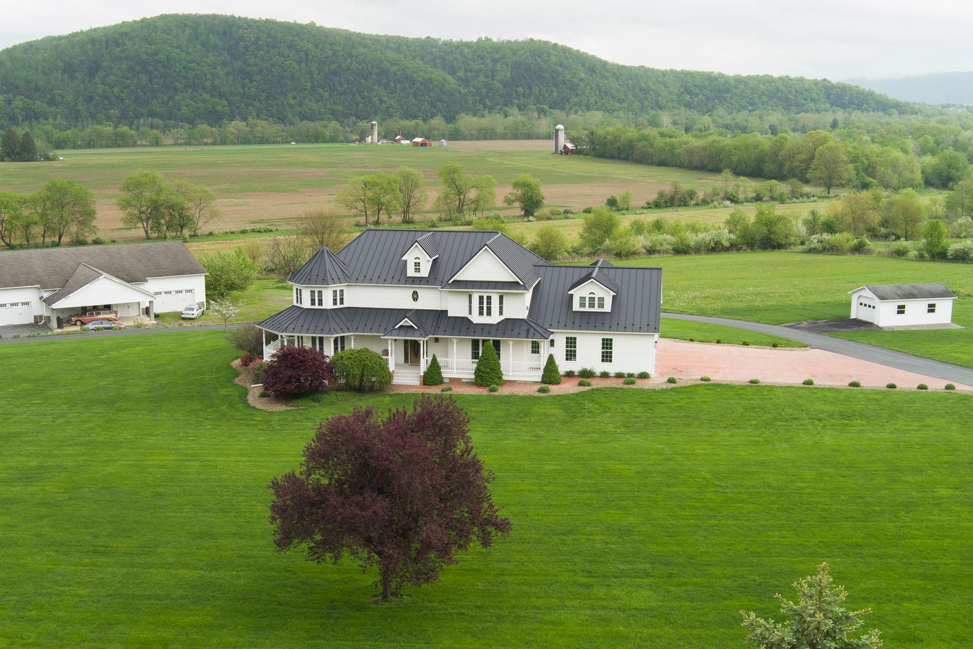 White house with black roof on a green lawn, surrounded by fields and a mountain range.