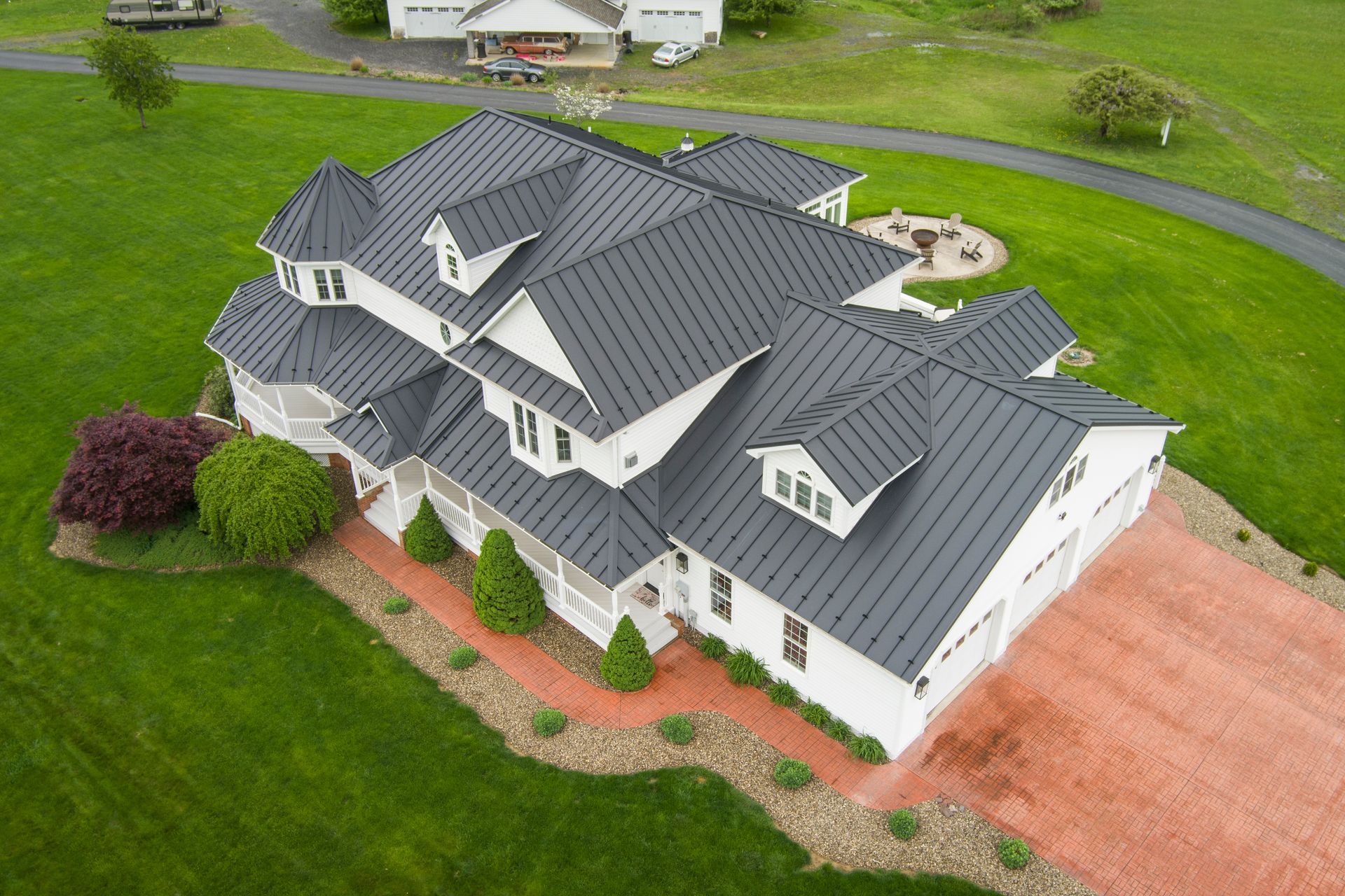 White house with dark gray metal roof, on a green lawn, red brick driveway.