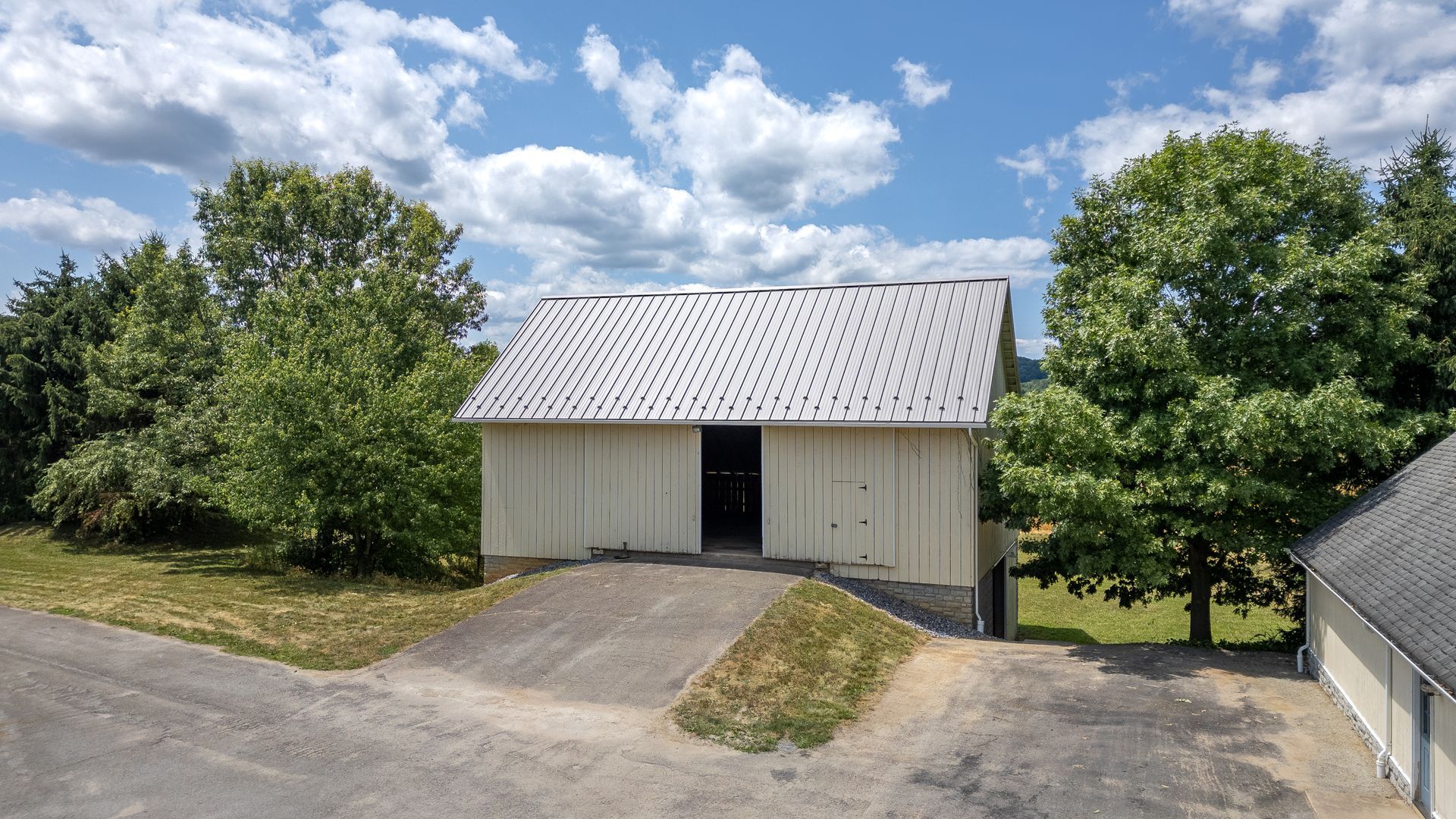 A white barn with a grey roof and open door, trees and blue sky in the background.