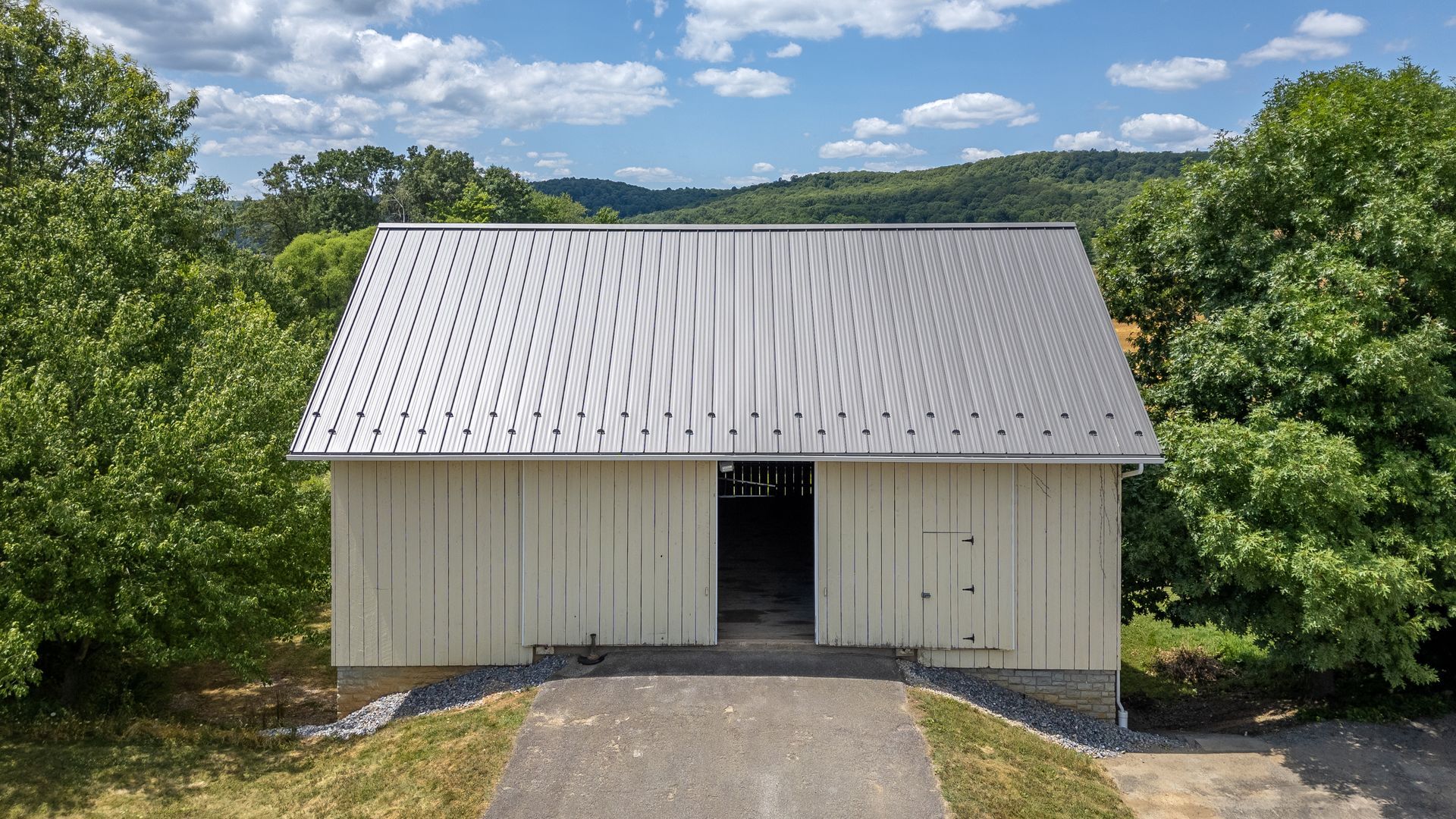 White barn with metal roof, open doors, and a paved driveway, surrounded by trees under a blue sky.