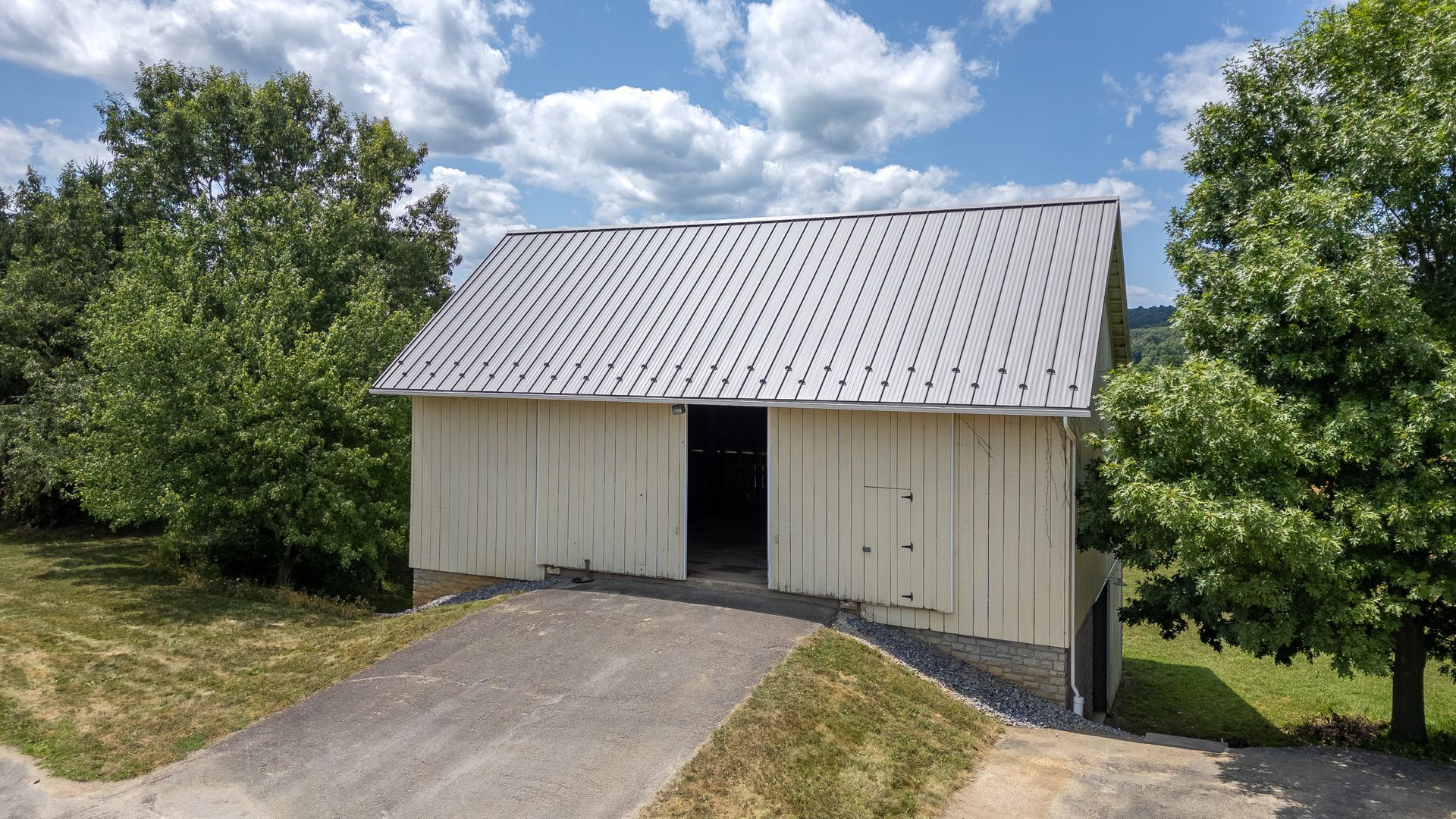 White barn with a metal roof, in front of a road with trees in the background.