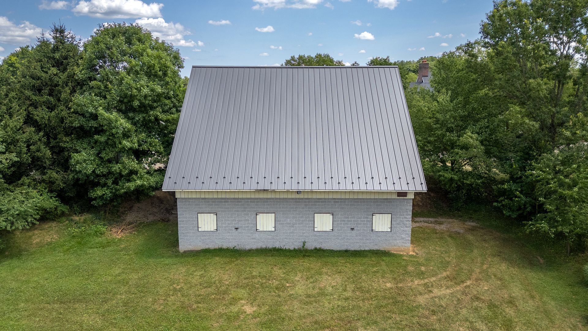 A gray-walled building with a metal roof sits in a grassy area, surrounded by trees, under a blue sky.
