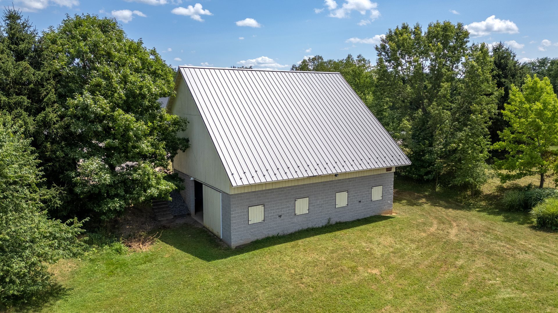 Barn with a gray exterior and a silver corrugated metal roof, surrounded by green trees and grass.