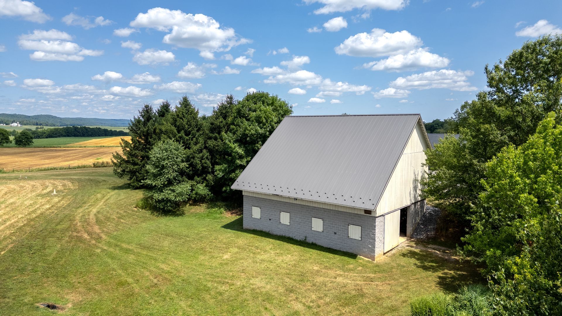 Barn in a field with a gray roof on a sunny day, with trees and a distant harvested field.
